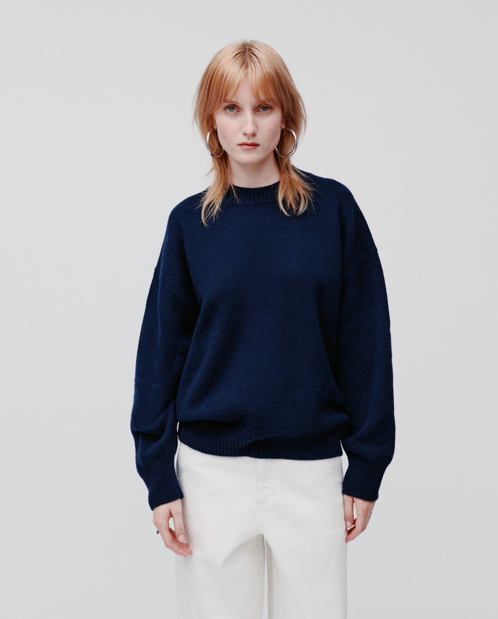 A woman with straight reddish-blonde hair wears the IVY OAK KASSIDY Jumper, a cozy oversized navy cashmere piece, paired with white pants against a plain background.