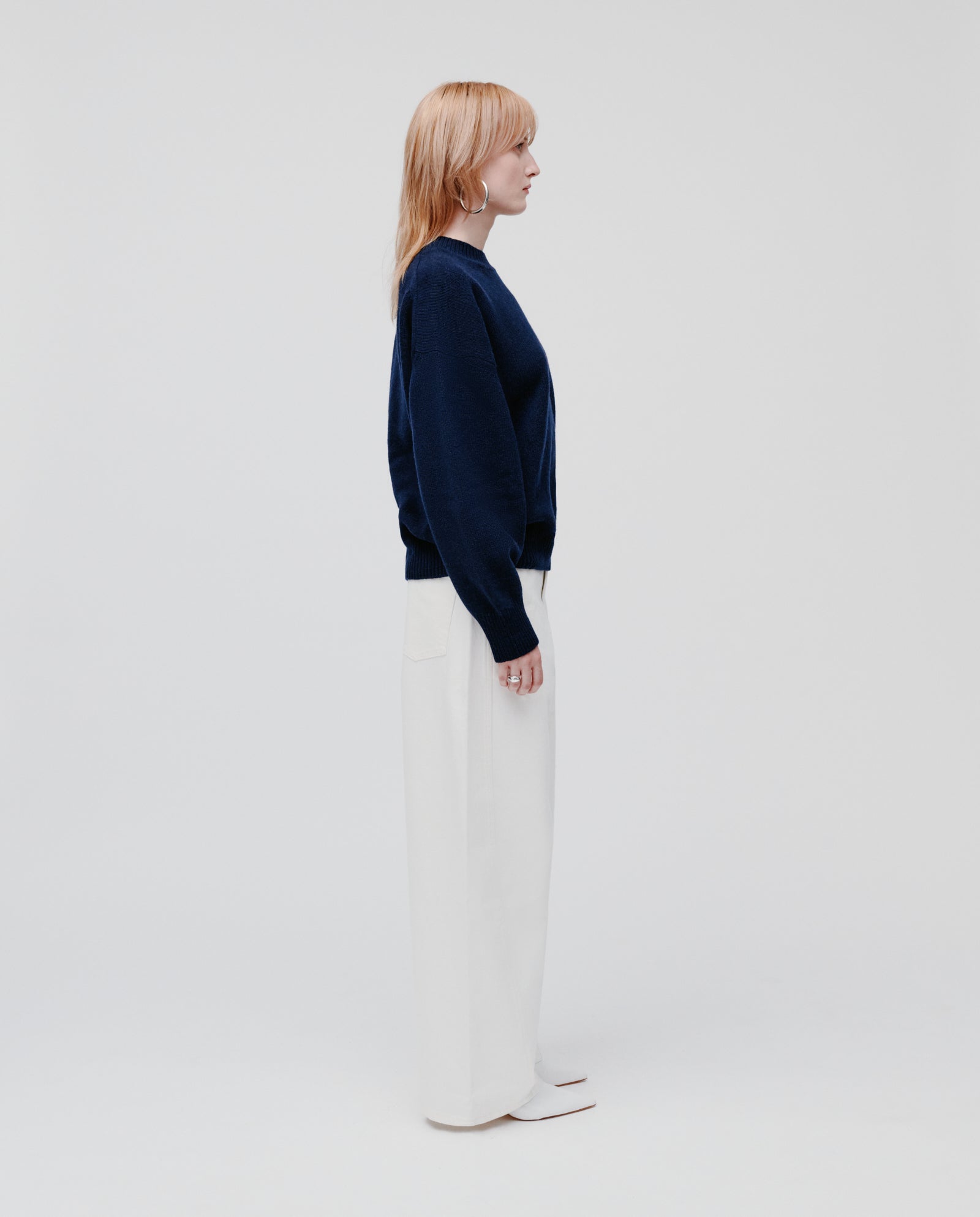 A person stands in profile against a plain light background, wearing the IVY OAK KASSIDY Jumper in cozy navy cashmere with an oversized fit, paired with white wide-leg pants.