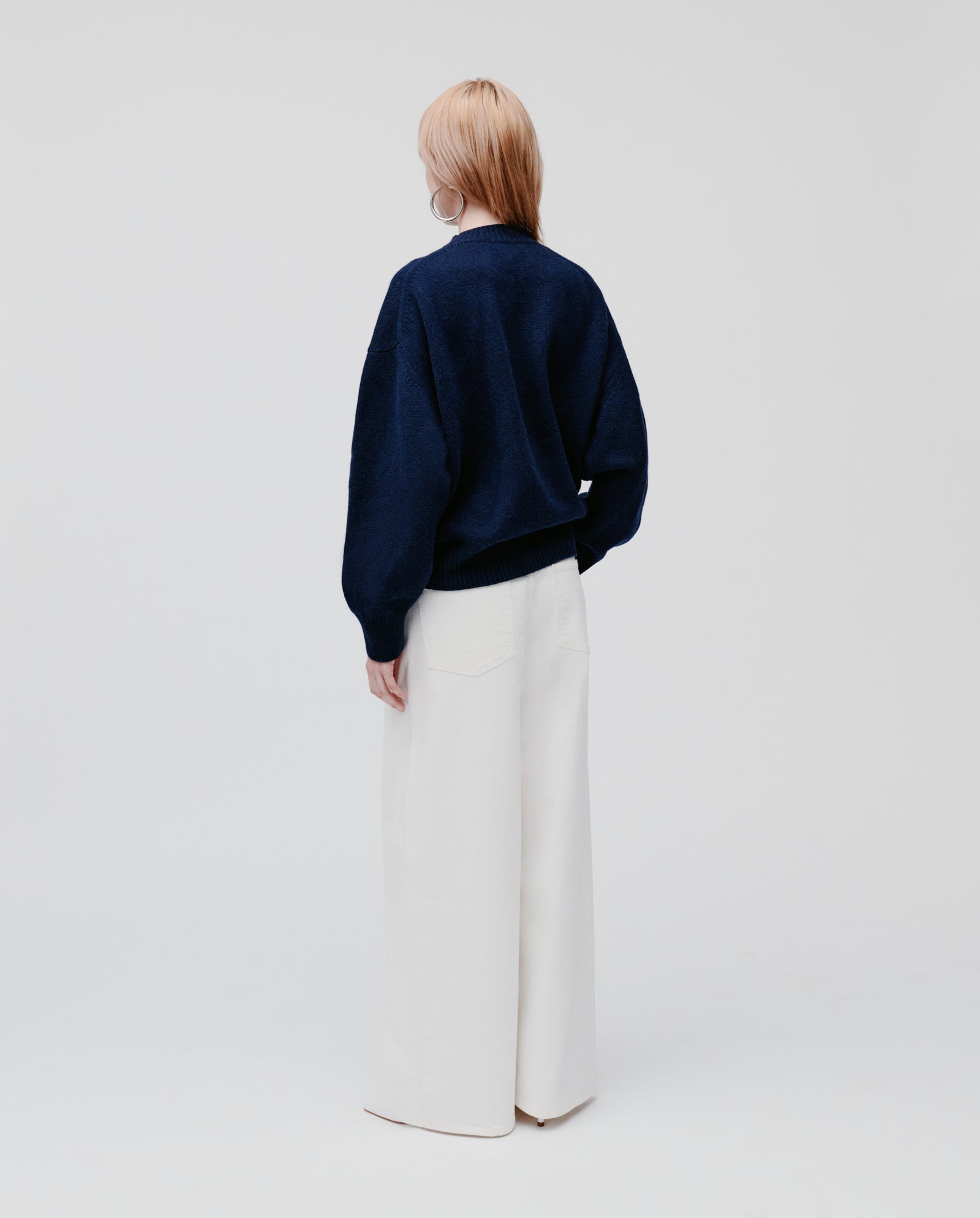 A person stands with their back to the camera, wearing the IVY OAK KASSIDY Jumper in cozy navy cashmere with an oversized fit and wide-leg white pants, set against a plain background.