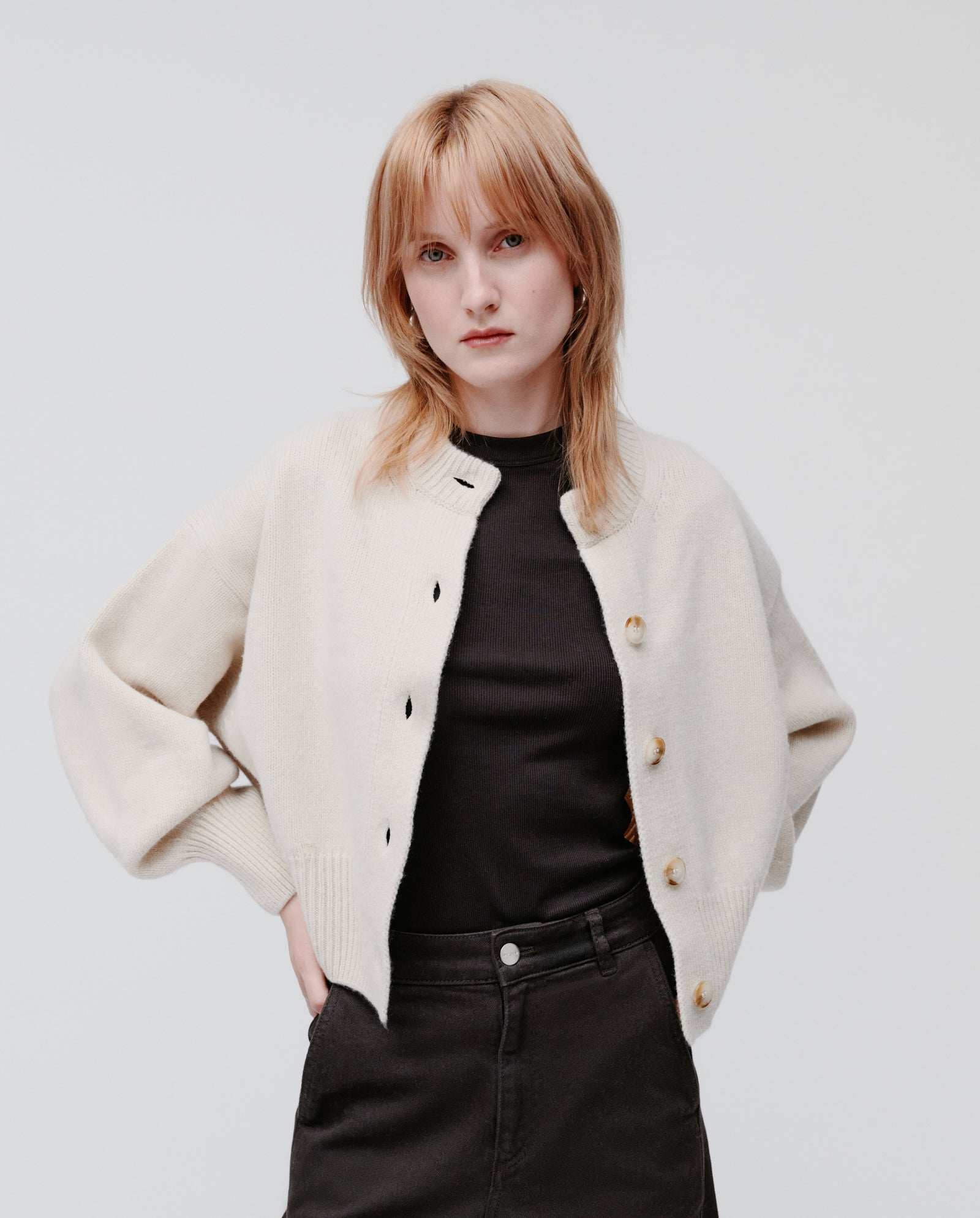 A woman with straight blonde hair wears the IVY OAK KARMA Cardigan—an oversized cream cashmere-blend style with wide sleeves—over a black shirt and pants, standing against a plain background.
