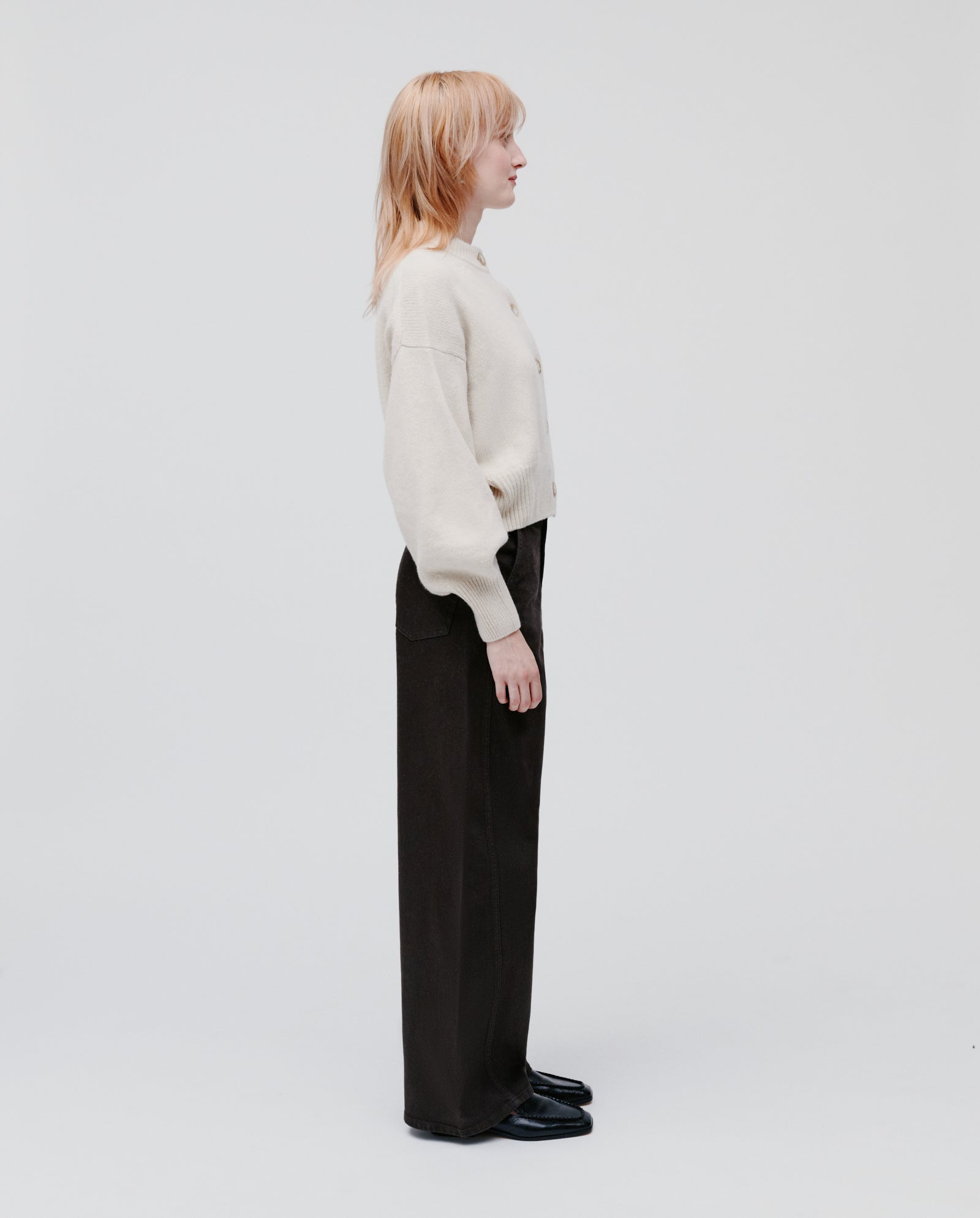 Person with shoulder-length light hair stands in profile, wearing the IVY OAK KARMA Cardigan in cream with wide sleeves and wide black pants.