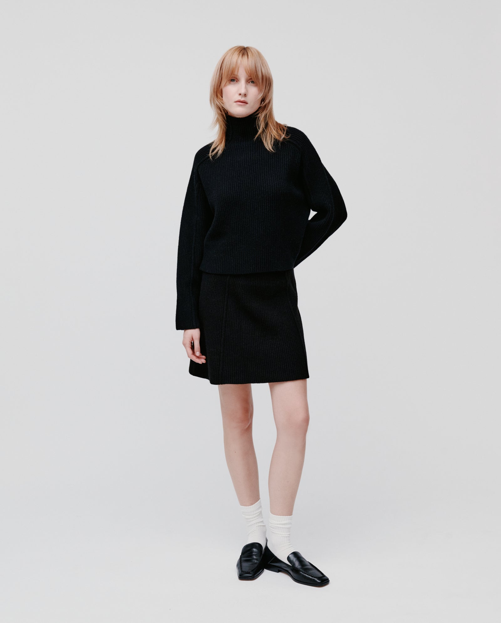 Woman in the IVY OAK KEEVA Jumper, an oversized black rib knit sweater, paired with a skirt, white socks, and black loafers stands against a white background.