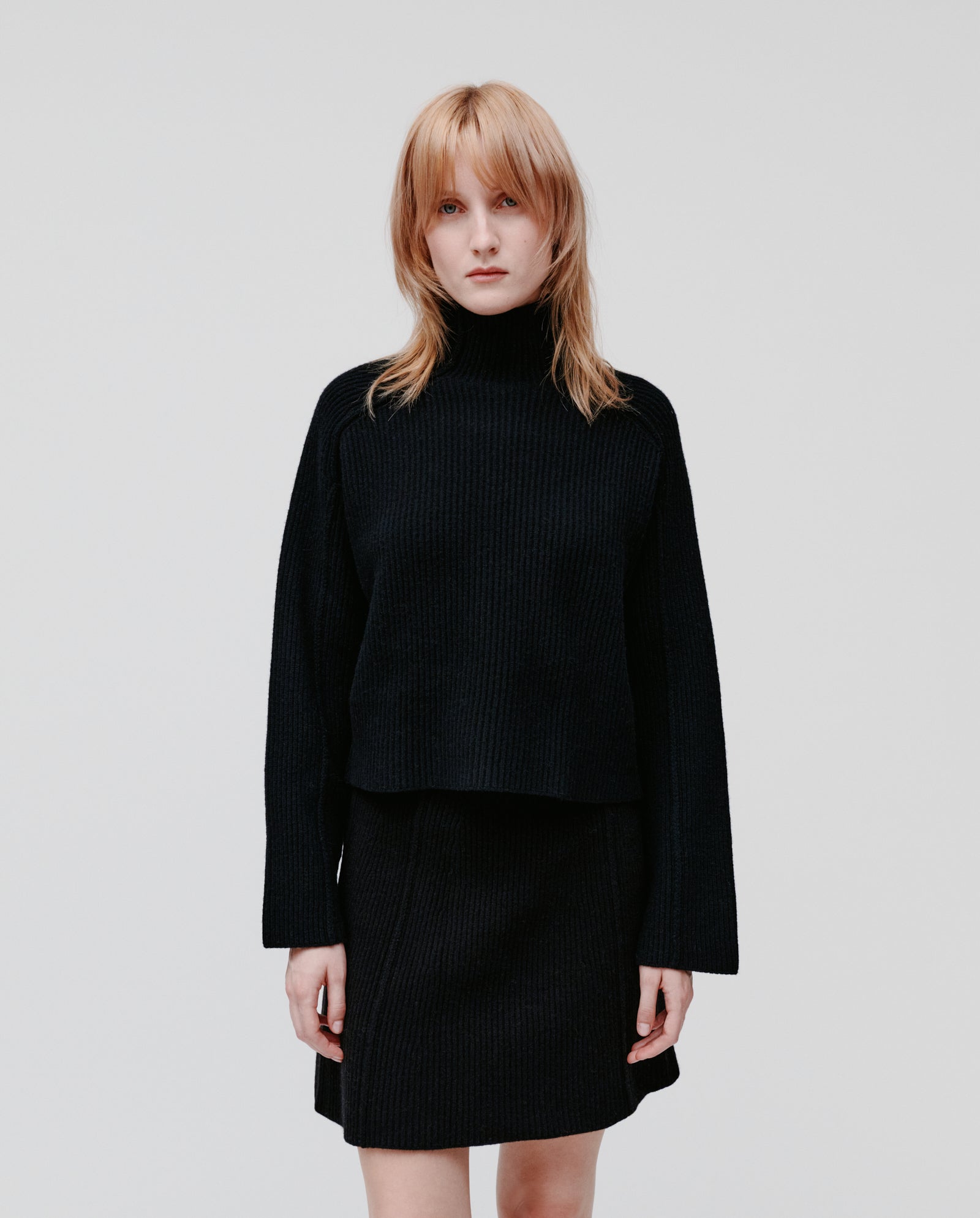 A woman wears the IVY OAK KEEVA Jumper, an oversized rib knit sweater, paired with a skirt, standing against a plain white background.