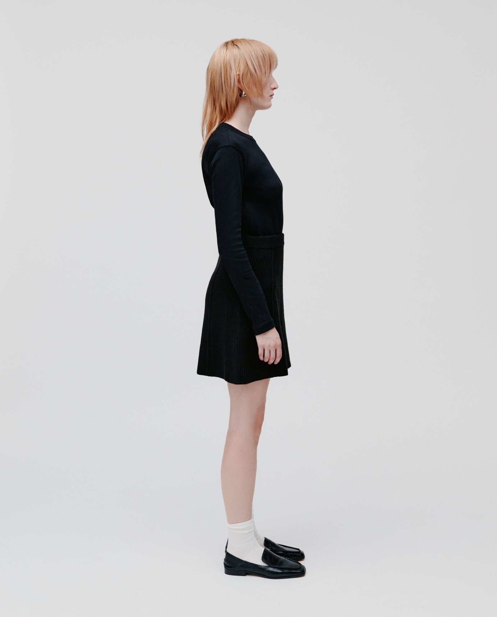 A woman with light hair stands in profile, dressed in a black top and the IVY OAK KEENA Skirt, paired with white socks and black shoes.