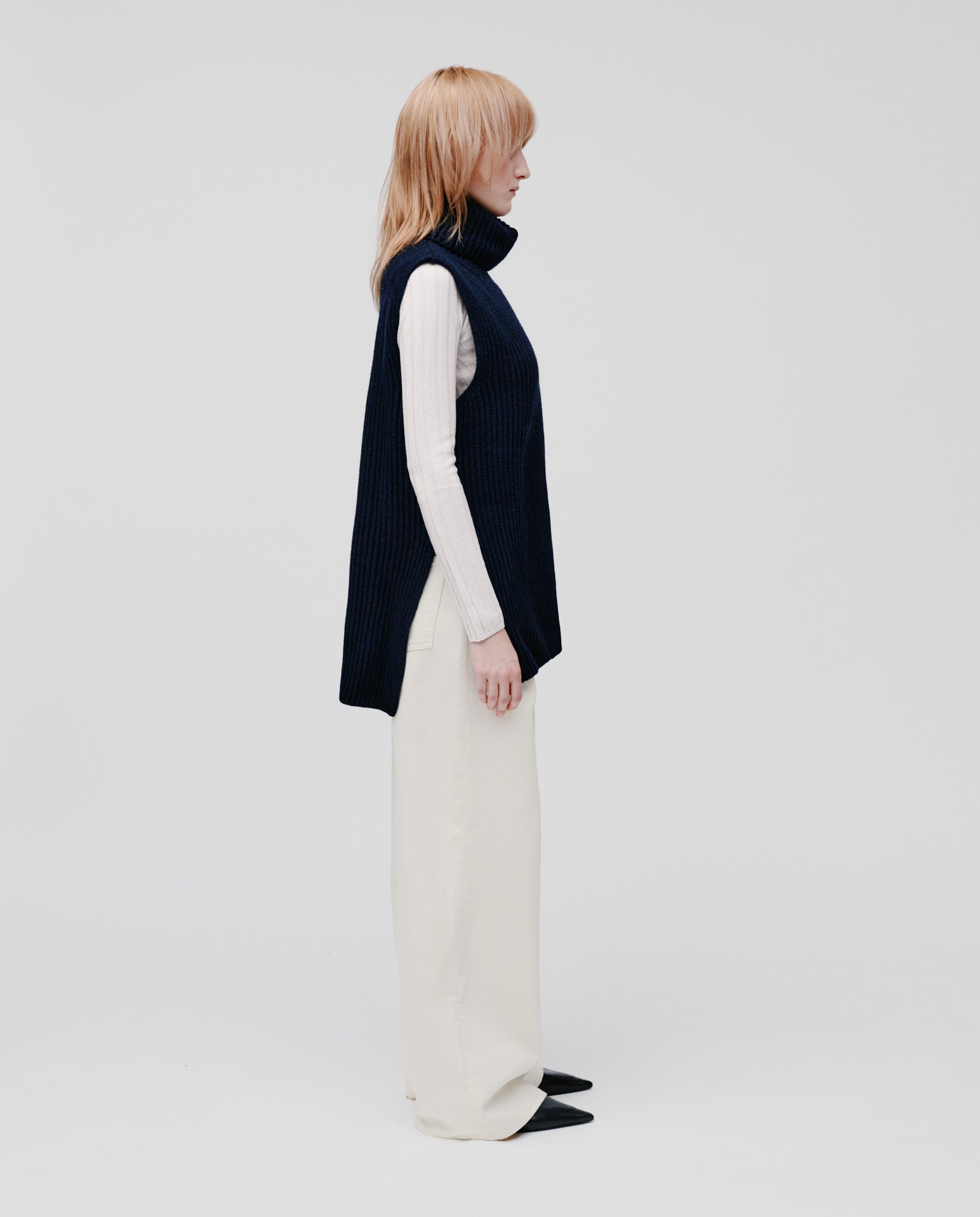 A person in profile wears the IVY OAK KYOMI Pullunder layered over a light turtleneck, paired with wide-leg cream pants.