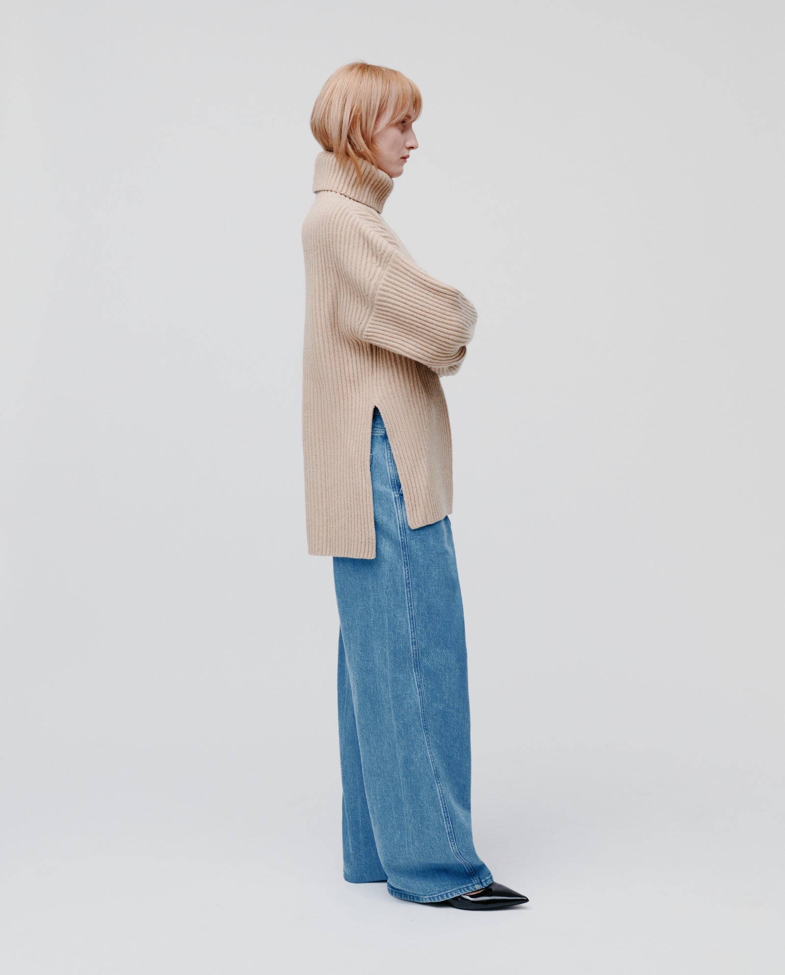 Model wearing the IVY OAK KYOMI ANN Jumper in beige and wide-leg blue jeans, standing sideways against a plain background.
