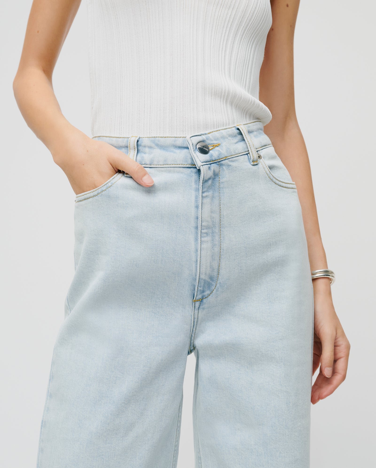Person wearing a white ribbed tank top and light blue high-waisted IVY OAK PIXIE Trousers with wide legs, one hand casually tucked in a patched pocket.