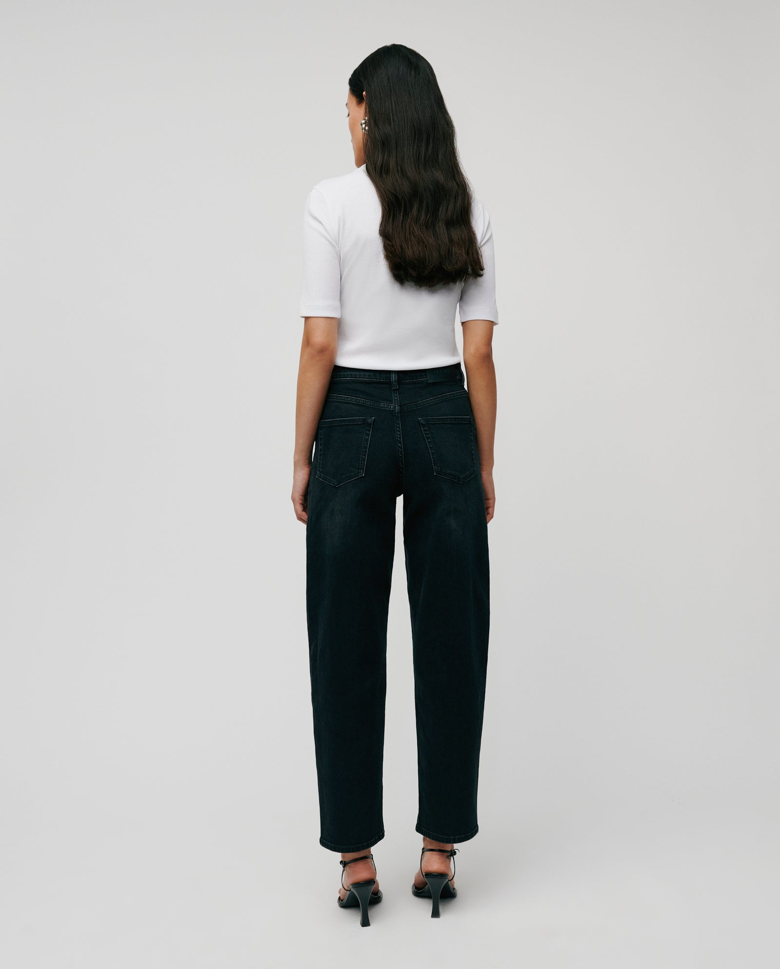 A woman stands facing away, wearing a white shirt and IVY OAK PETRA Trousers with patched pockets and a barrel-leg silhouette, paired with heels against a plain background.