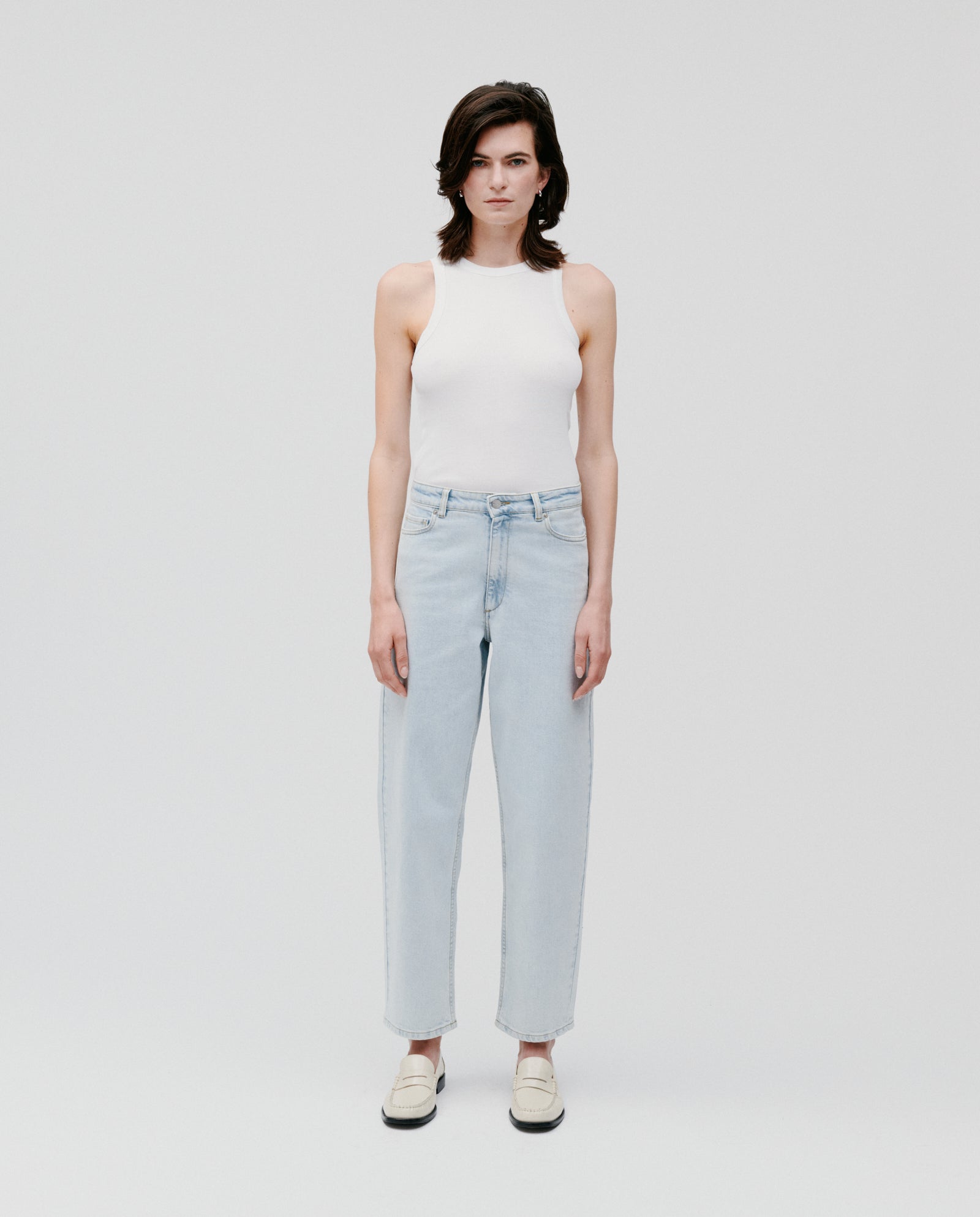 Person stands against a plain background, wearing a white tank top, white shoes, and IVY OAKs PETRA Trousers—light blue barrel leg jeans with patched pockets.