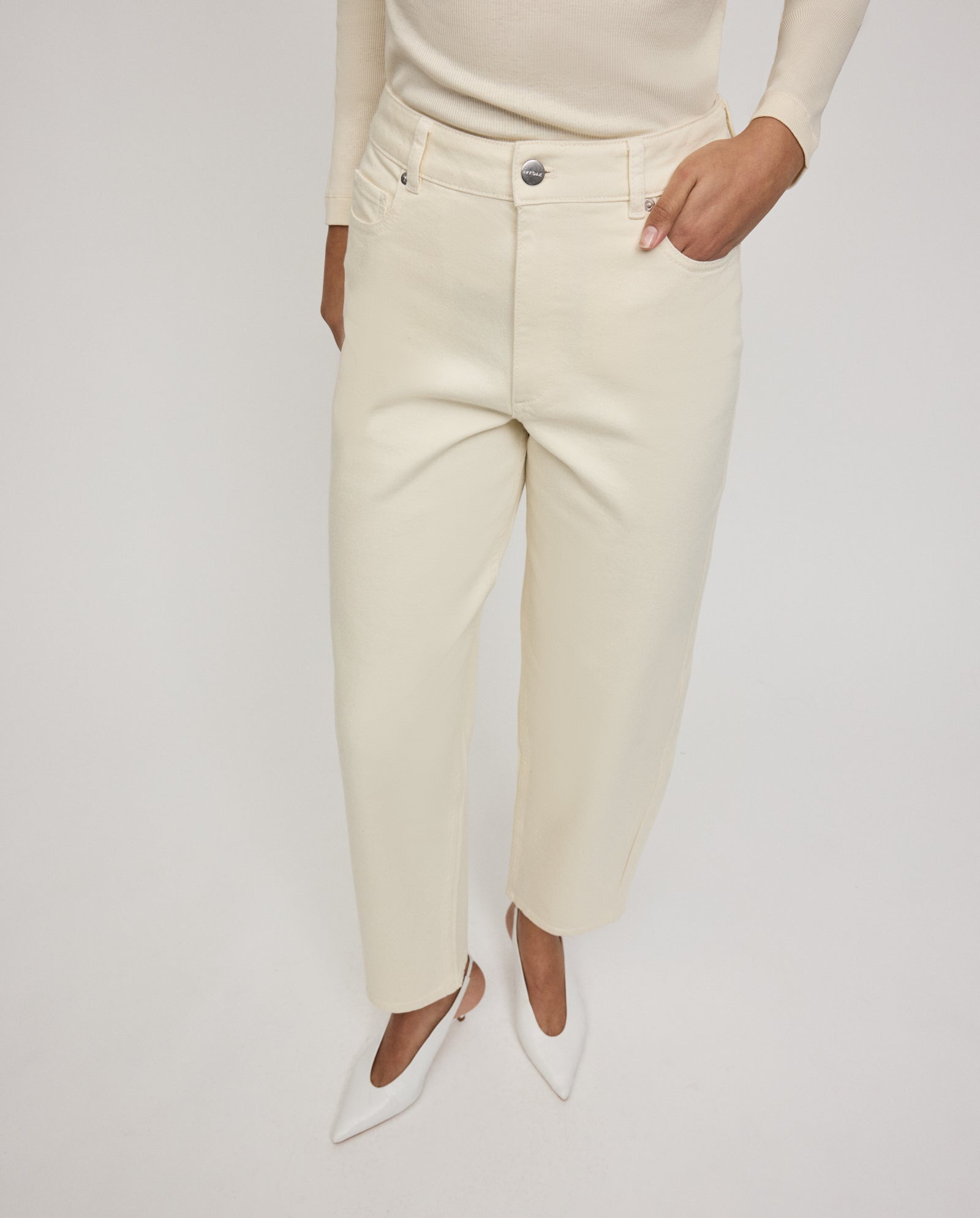 Model wearing IVY OAK PETRA Trousers in off-white barrel-leg style with patch pockets, paired with white pointed heels and a long-sleeve top, hand in pocket, standing against a white background.