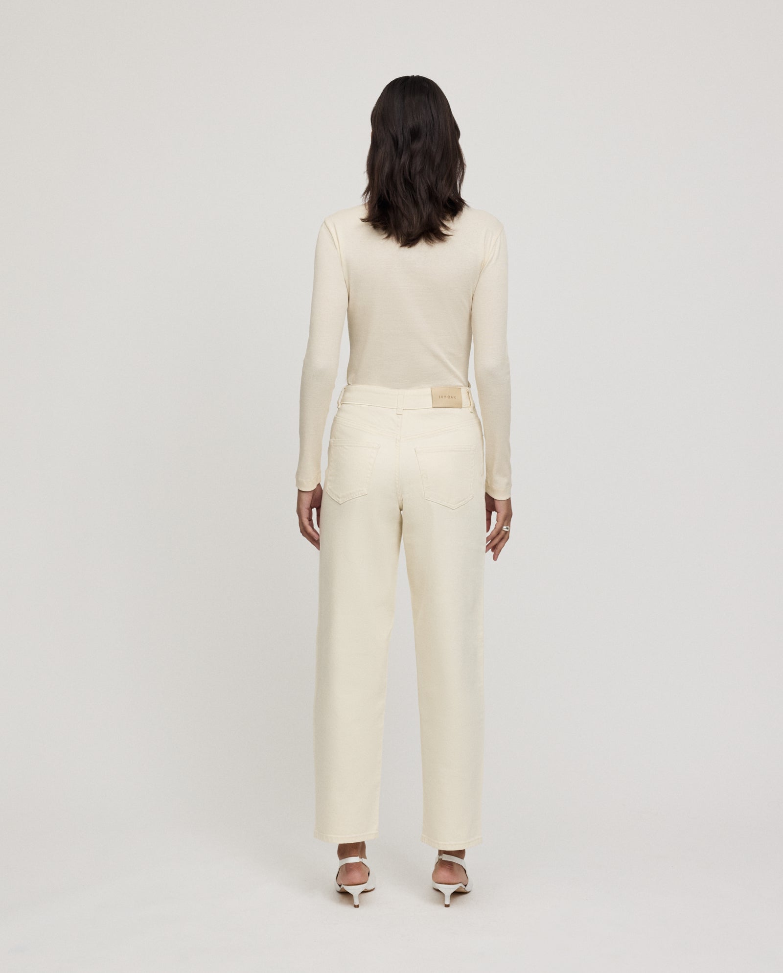 Woman standing facing away, wearing a cream long-sleeve top, IVY OAK PETRA cream barrel-leg trousers with patch pockets, and white heeled sandals.