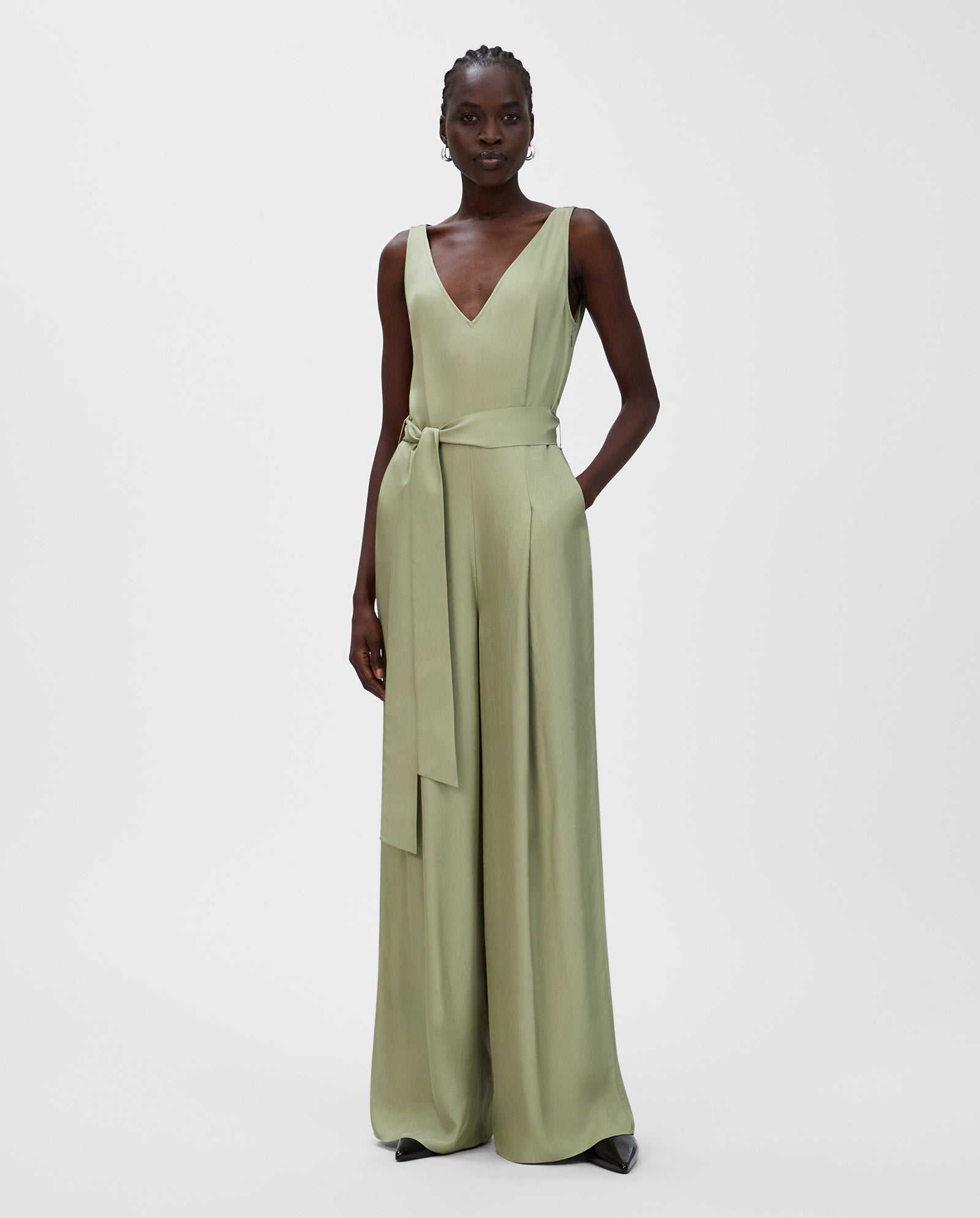 A woman wears the IVY OAK PATRICIA Jumpsuit—a sleeveless, light green, V-neck design with a waist tie and wide legs, made from soft Naia acetate—standing against a white background.