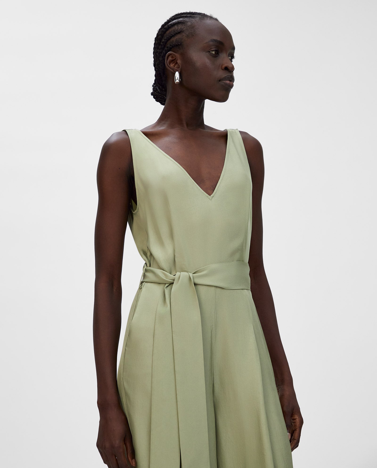 Woman wearing a light green IVY OAK PATRICIA Jumpsuit in Naia acetate, featuring wide legs and a waist tie, gazing to the side against a plain background.