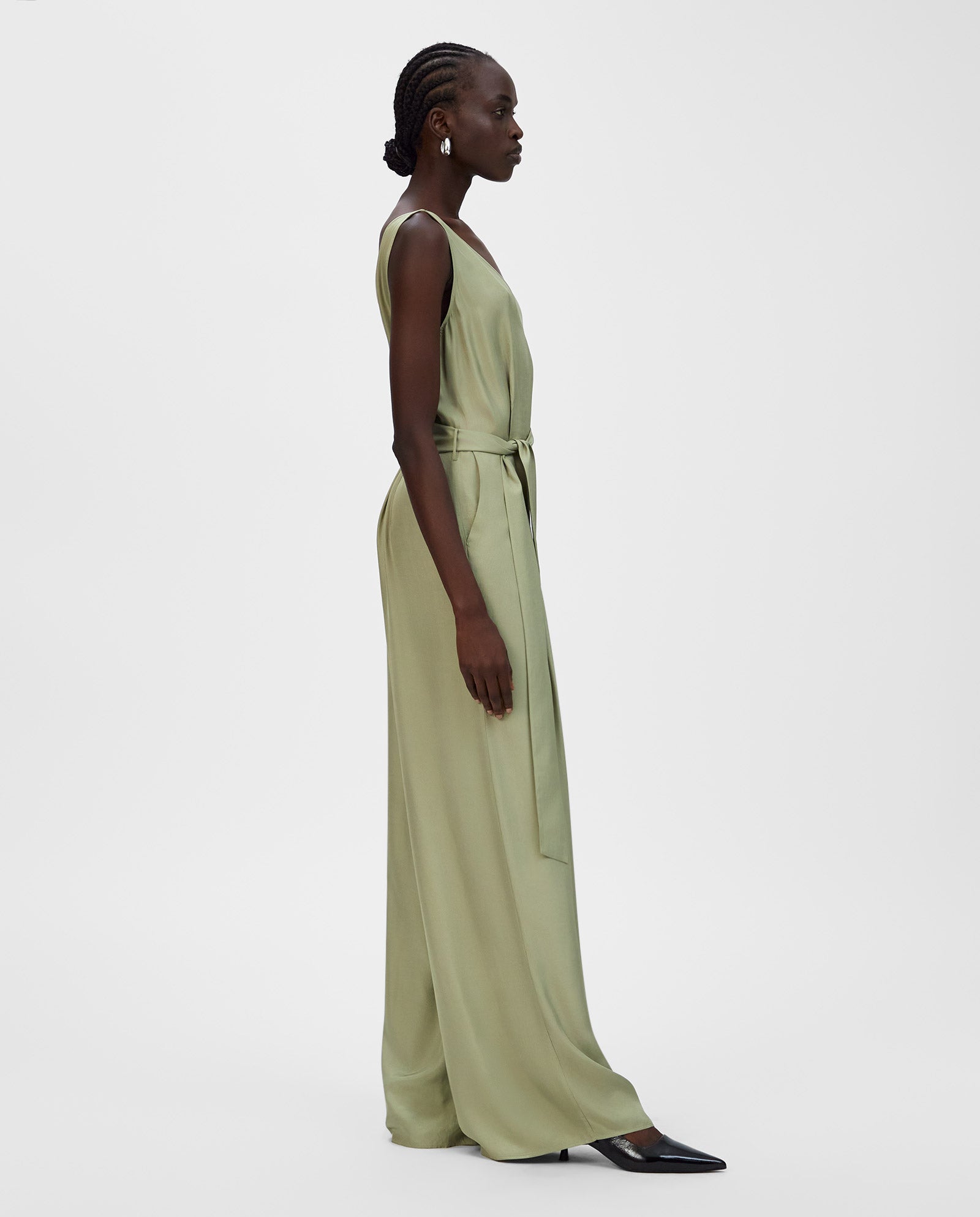 A woman in a light green IVY OAK PATRICIA Jumpsuit with wide legs and a belted waist, crafted from Naia acetate, pairs it with black heels and stands in profile against a white background.