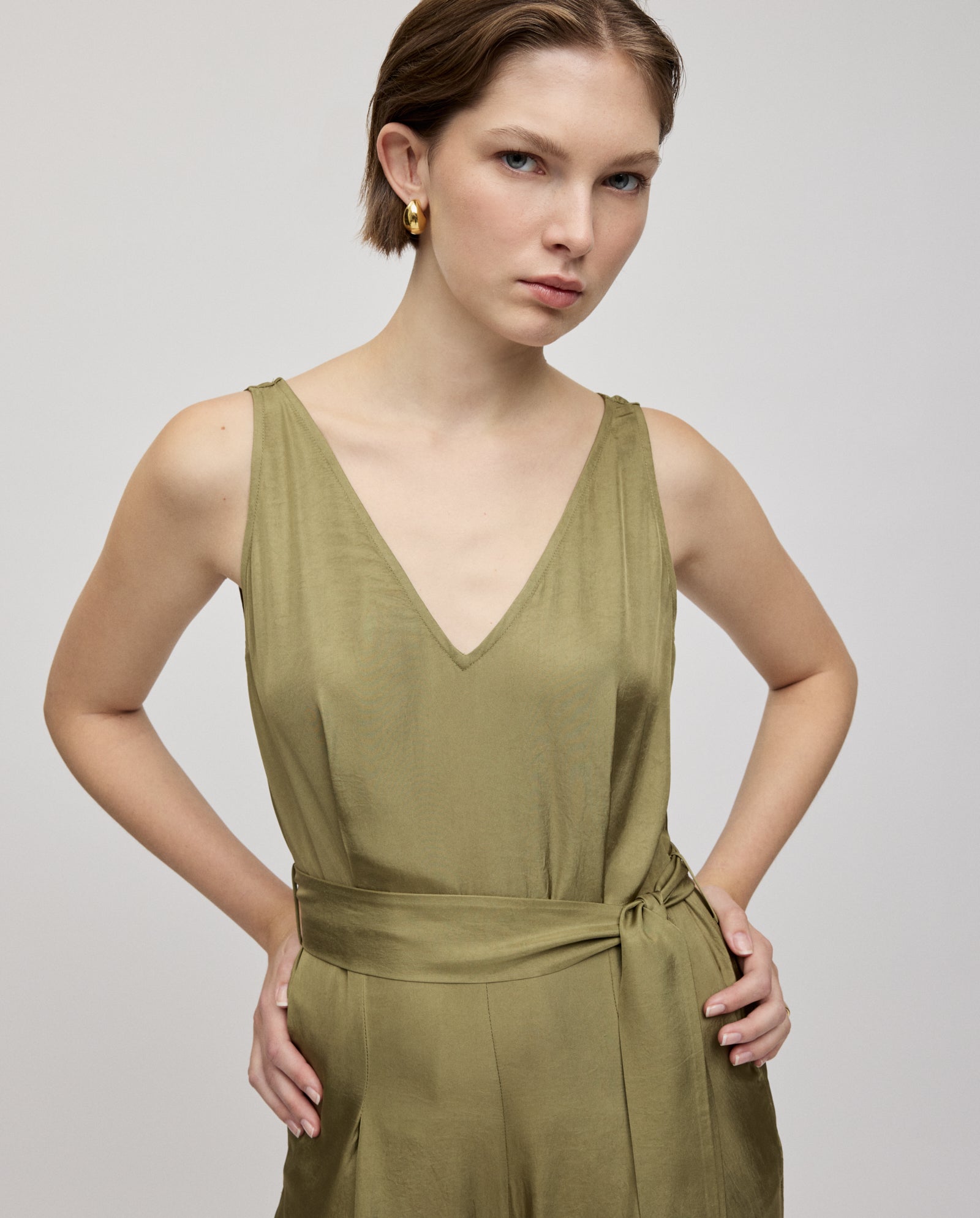 A woman stands with hands on hips, wearing the IVY OAK PATRICIA Jumpsuit in olive green Naia acetate, featuring a V-neck, sleeveless cut, and adjustable belt.