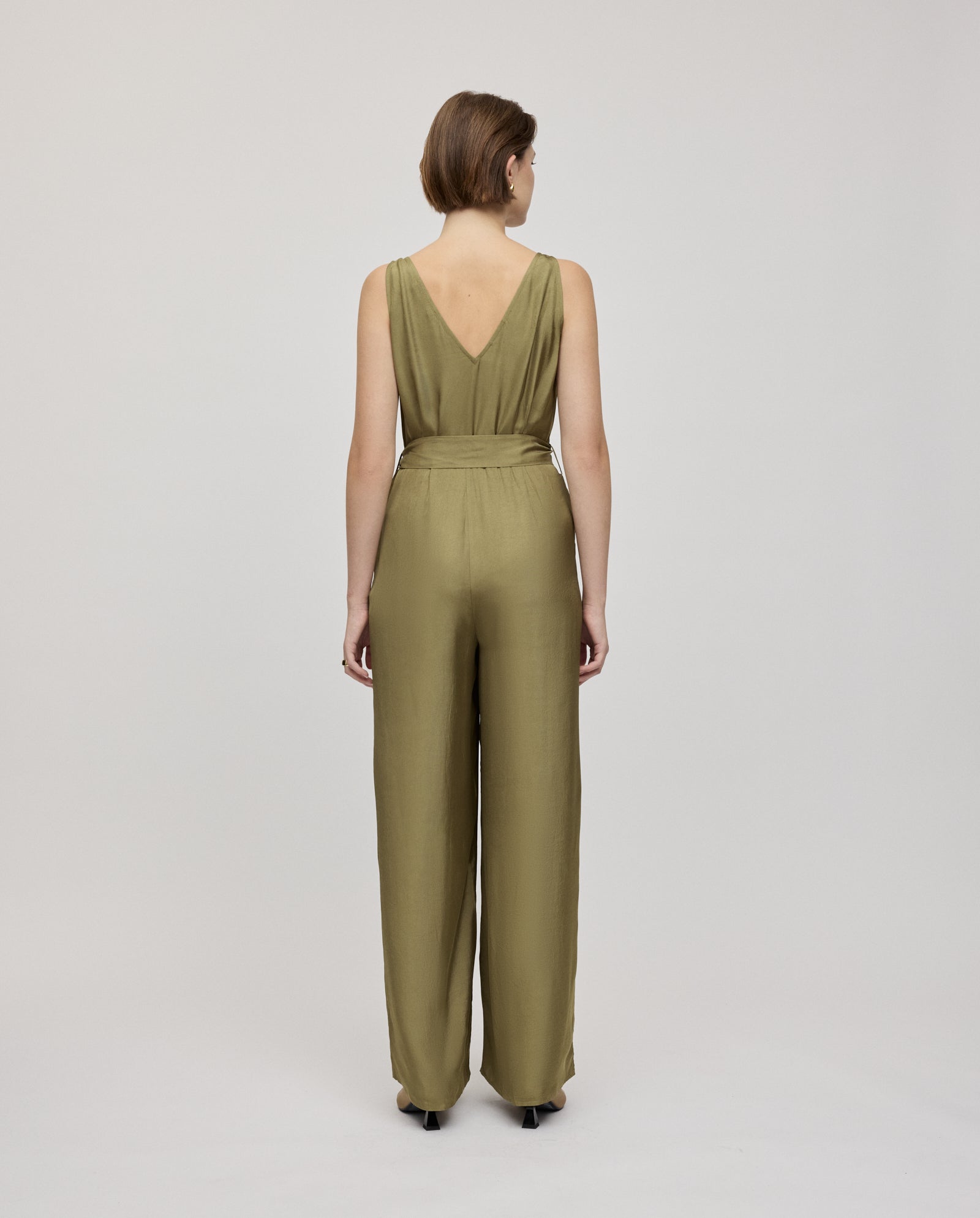 A woman stands facing away in an olive green PATRICIA Jumpsuit by IVY OAK, crafted from Naia acetate, featuring a deep V-back and adjustable belt.