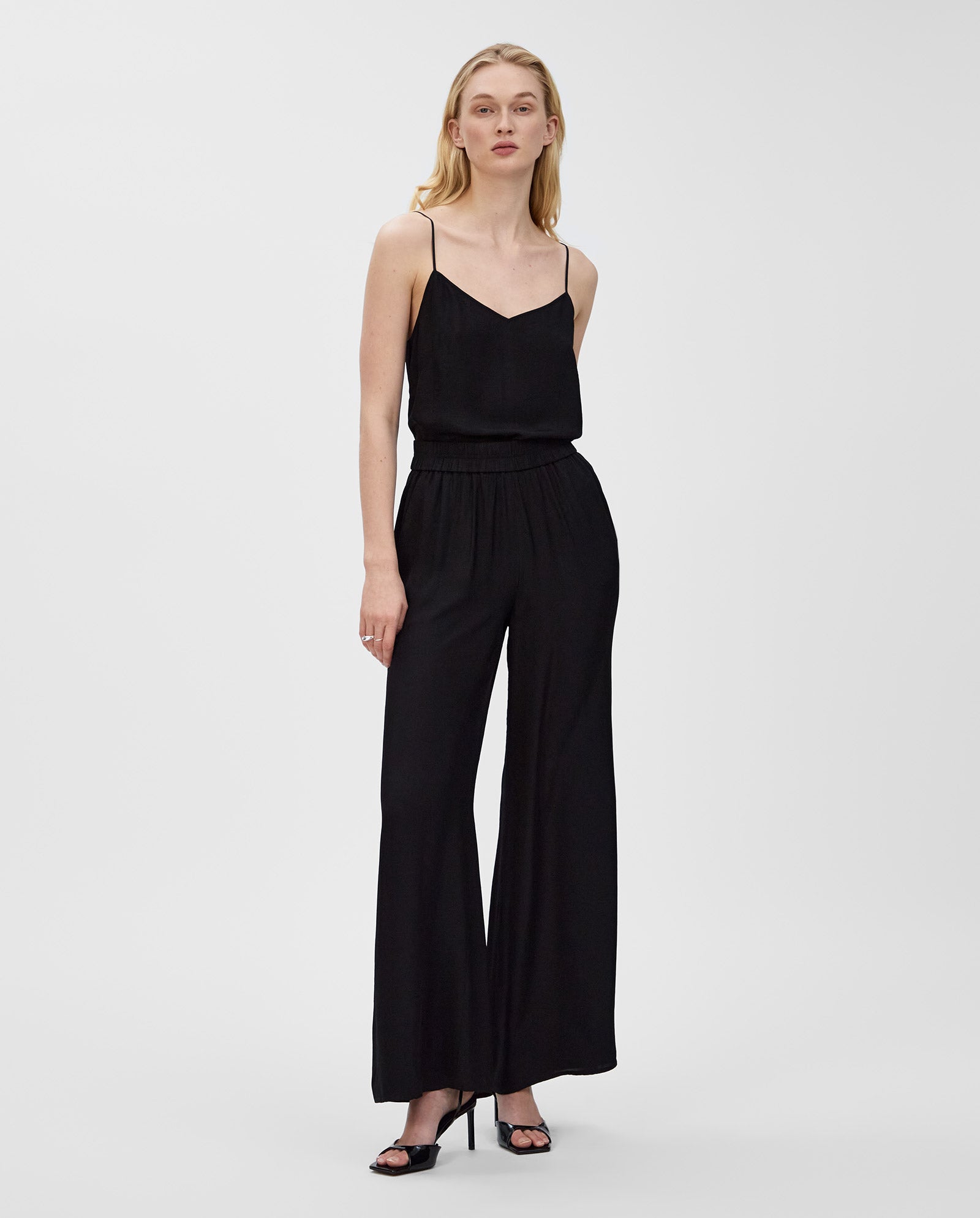 A woman wears IVY OAKs PIA ROSE Trousers, a black wide-leg style crafted from soft Naia acetate, standing against a plain background.