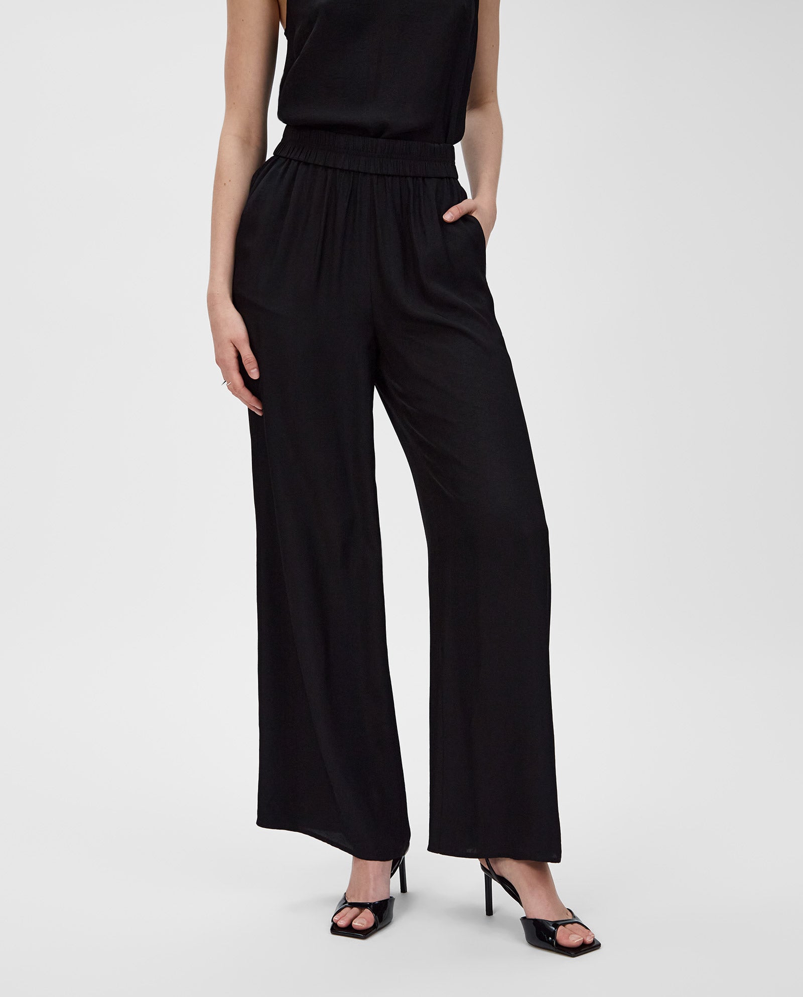 A woman wears IVY OAKs PIA ROSE Trousers, featuring wide legs and crafted from soft Naia acetate, styled with a sleeveless top and black heeled sandals against a white background.