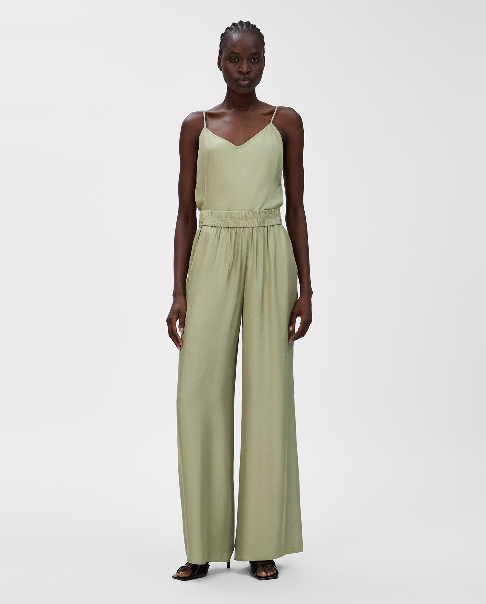 A woman wearing IVY OAK’s PIA ROSE Trousers—a light green, sleeveless, wide-leg jumpsuit with an elastic waistband crafted from soft Naia acetate—pairs them with black shoes against a plain background.