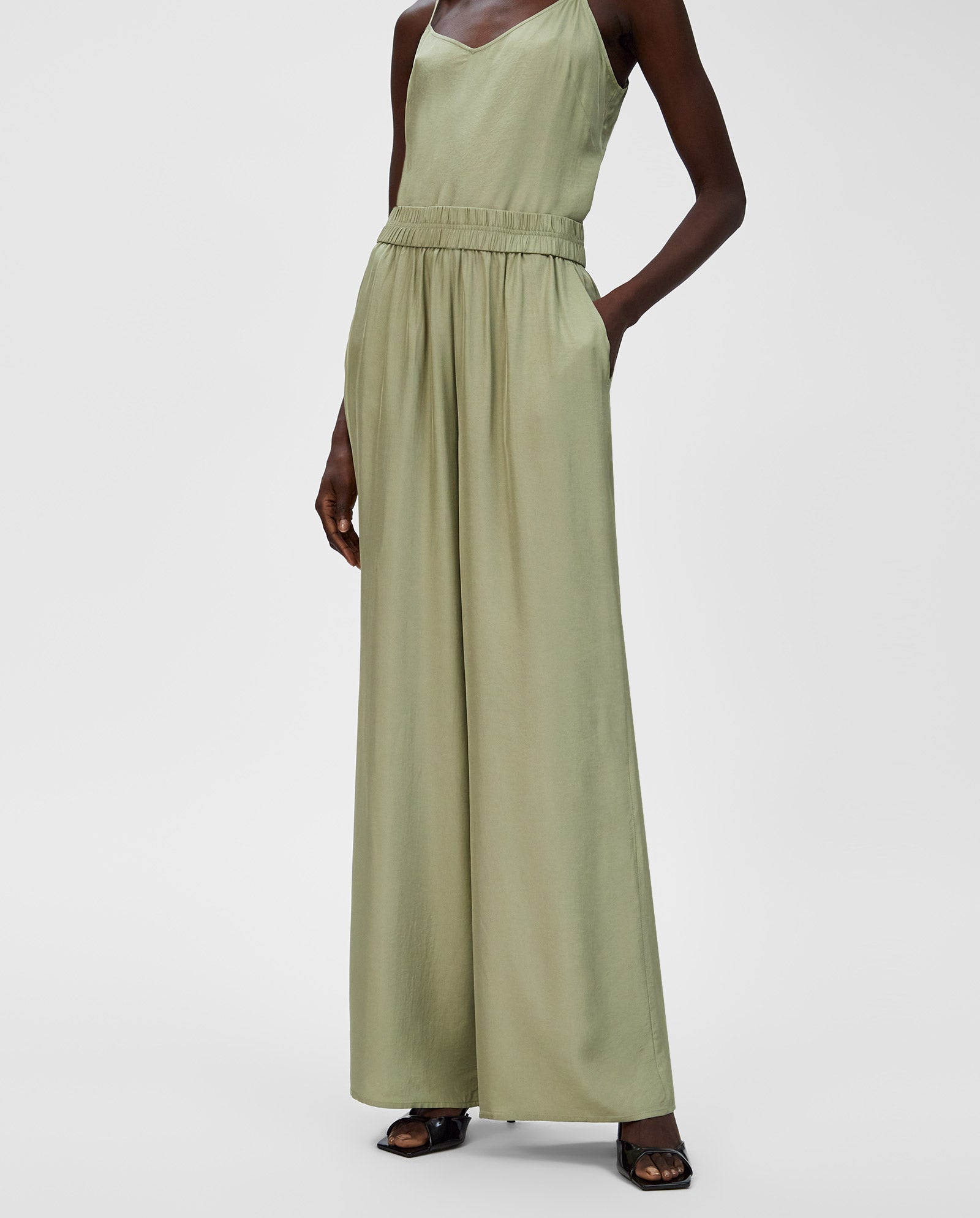 A woman in IVY OAKs PIA ROSE Trousers, a light green sleeveless jumpsuit with wide-leg Naia acetate pants, black sandals, and hands in pockets.