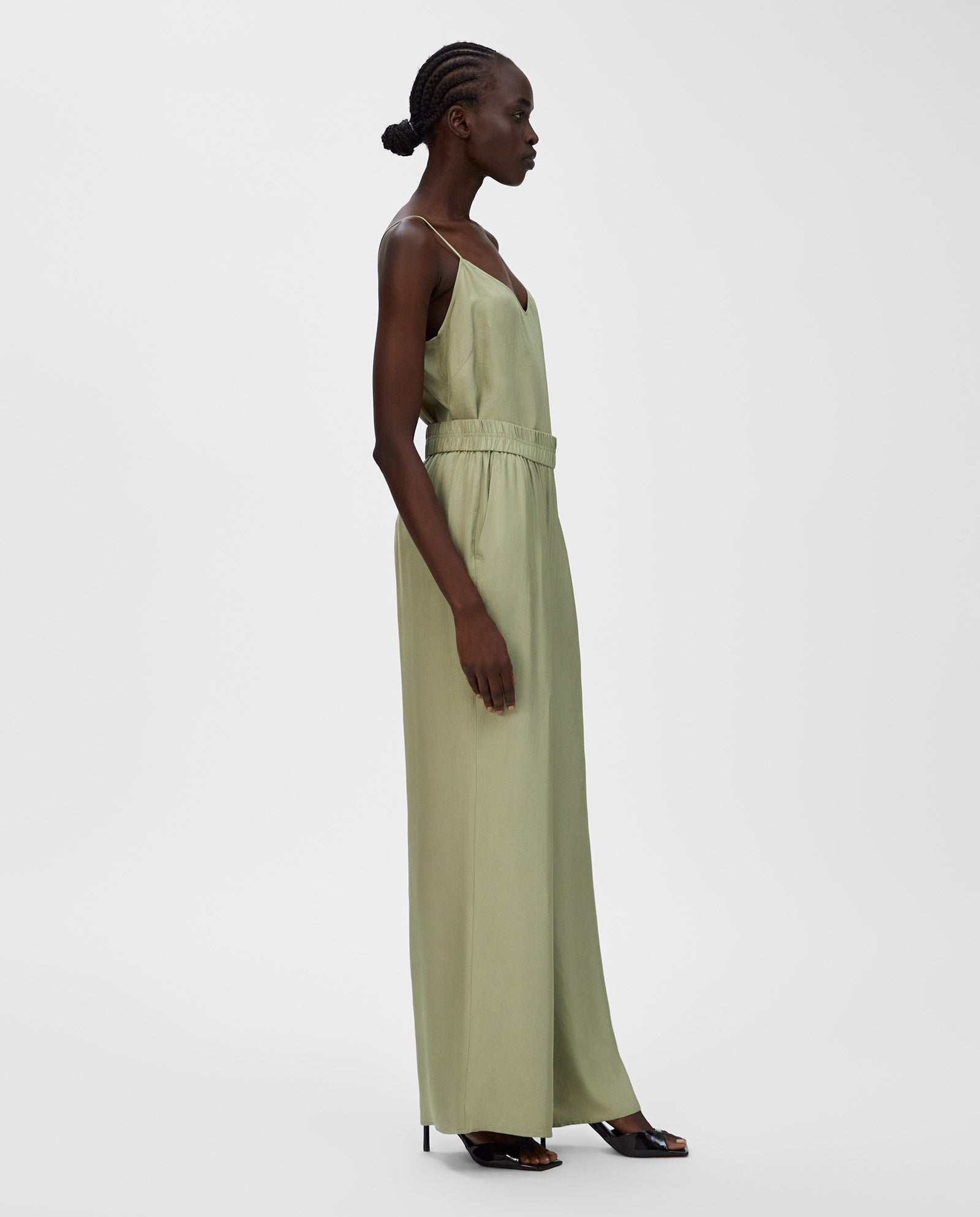 A woman in IVY OAK’s PIA ROSE Trousers—light green, wide-leg, sleeveless jumpsuit with an elastic waistband—stands sideways against a plain white background.