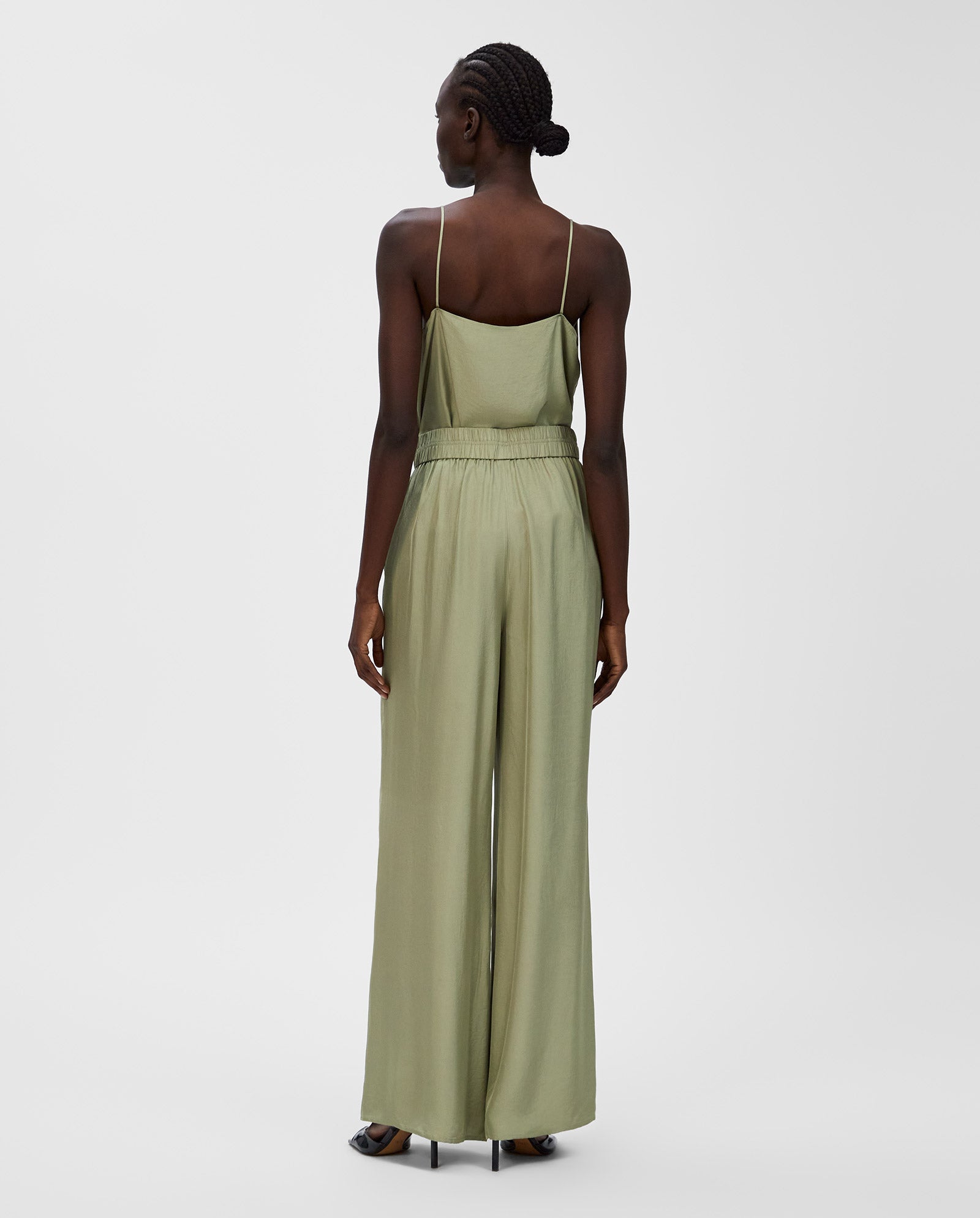A woman stands with her back to the camera in IVY OAK’s PIA ROSE sage green spaghetti strap jumpsuit featuring wide-leg trousers and an elastic waistband, set against a plain white background.