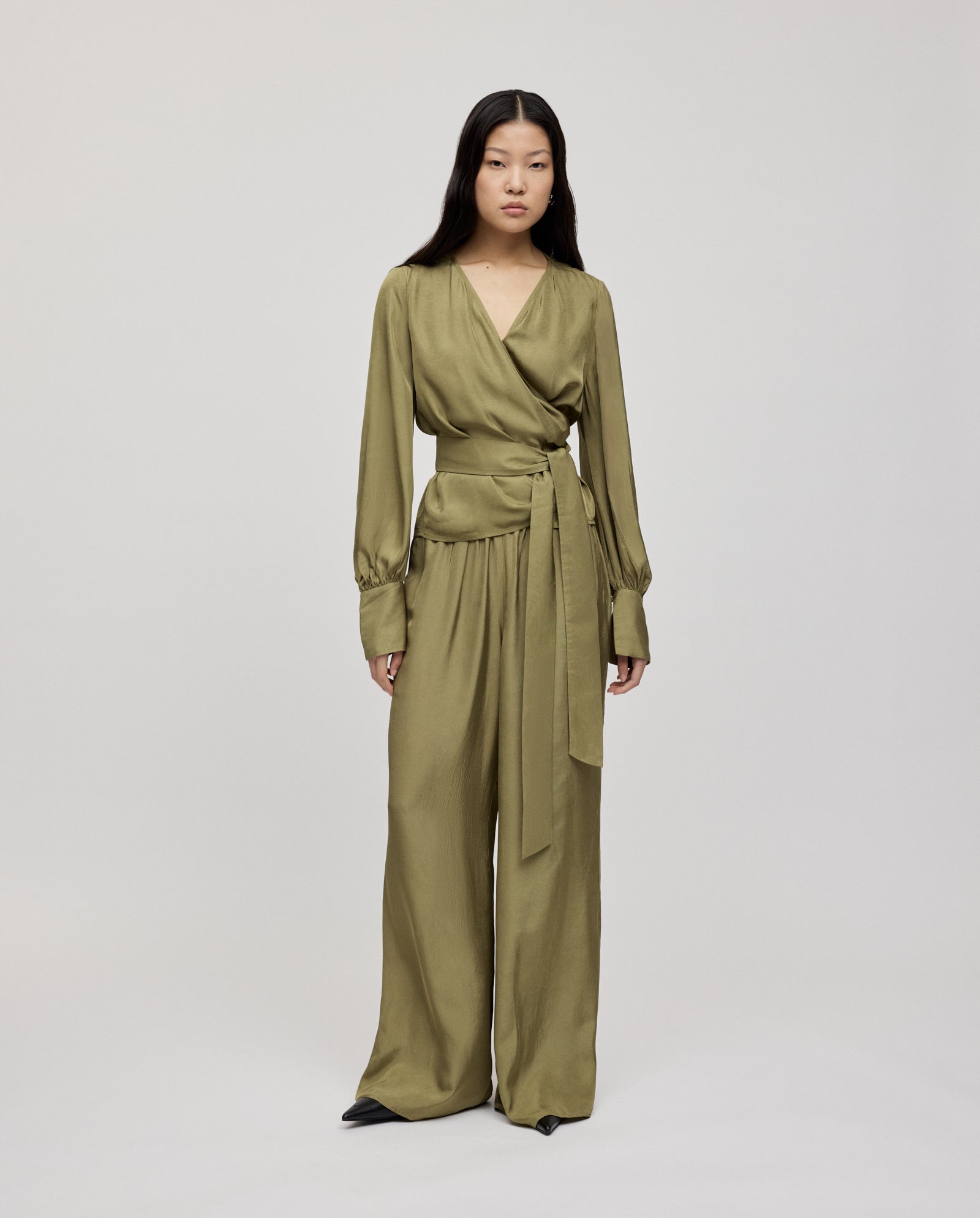 A woman models olive green satin IVY OAK PIA ROSE trousers, crafted from certified Naia acetate, paired with a coordinating wrap top against a plain background.