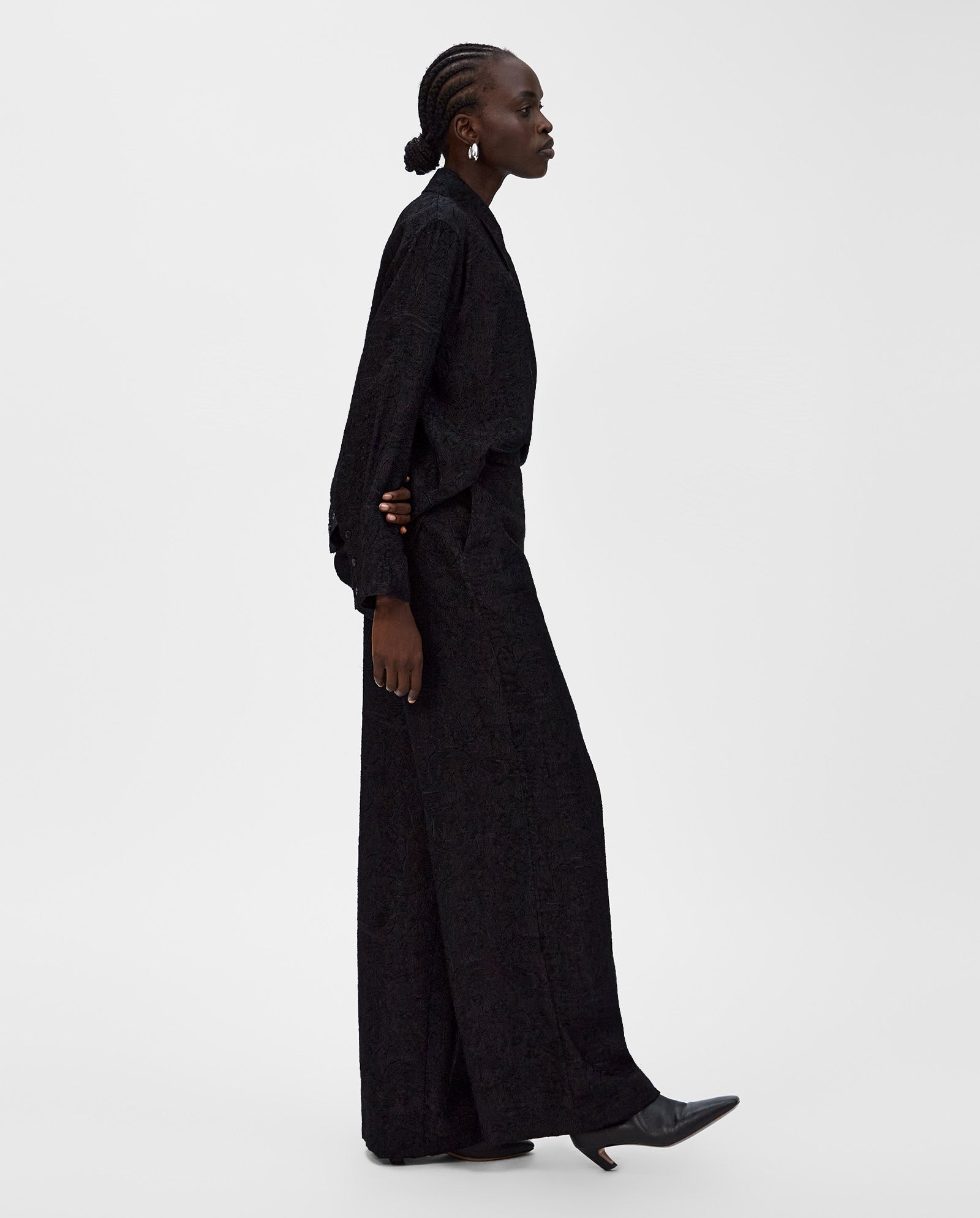 A person in a long black textured fil coupé outfit featuring IVY OAKs high-waisted PRIYA Trouser and heeled boots, walking against a plain white background.