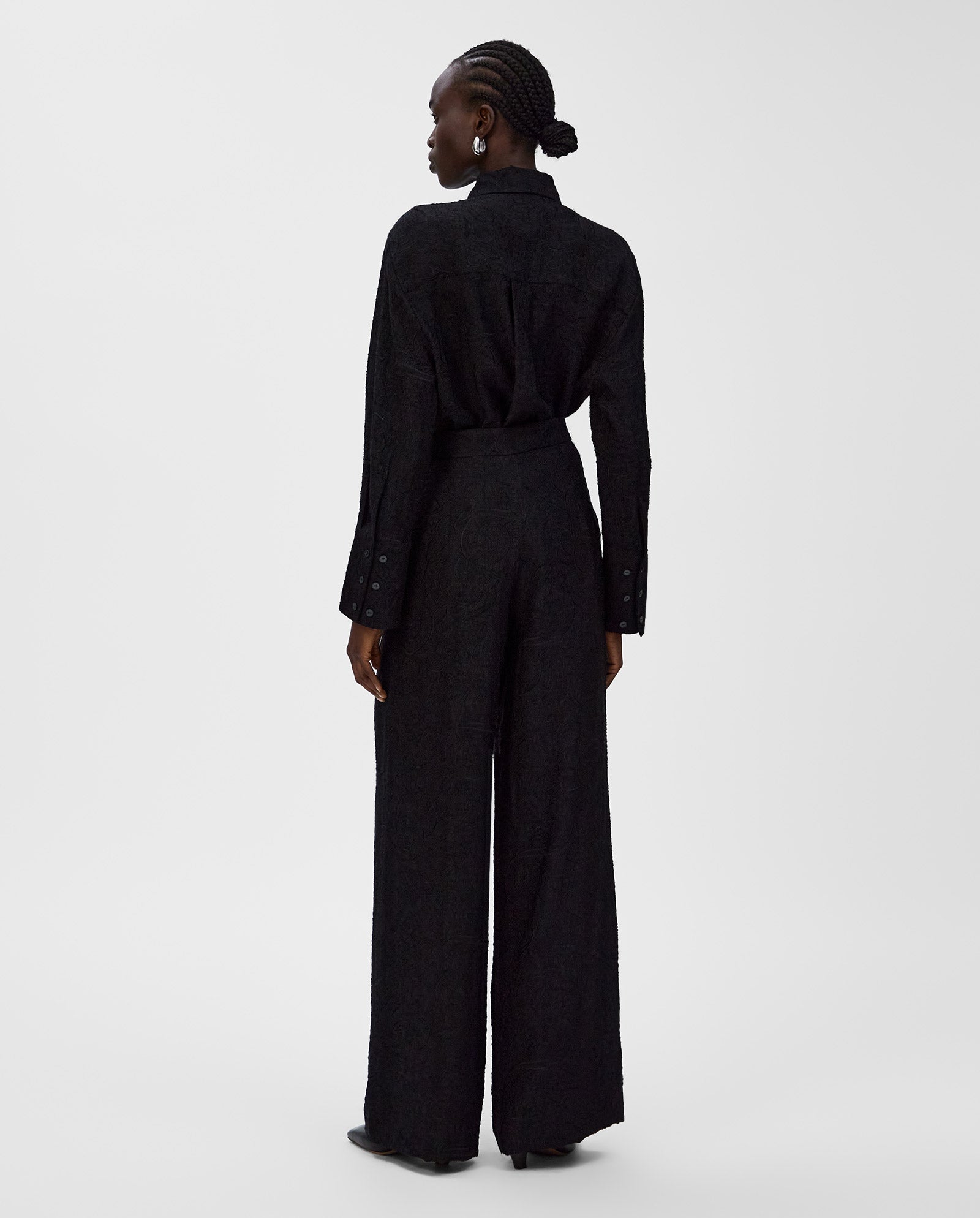 A woman stands with her back to the camera, wearing a black long-sleeve shirt and high-waisted IVY OAK PRIYA Trouser.