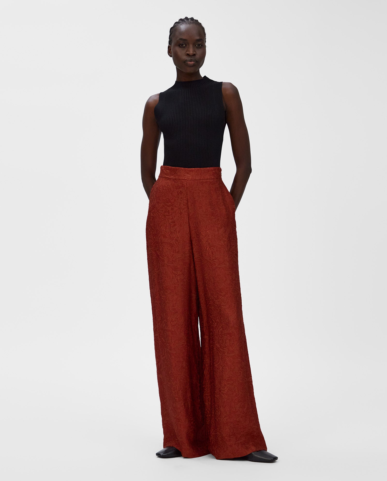 A woman wears a sleeveless black top with high-waisted, wide-leg IVY OAK PRIYA Trousers in rust red and black shoes.
