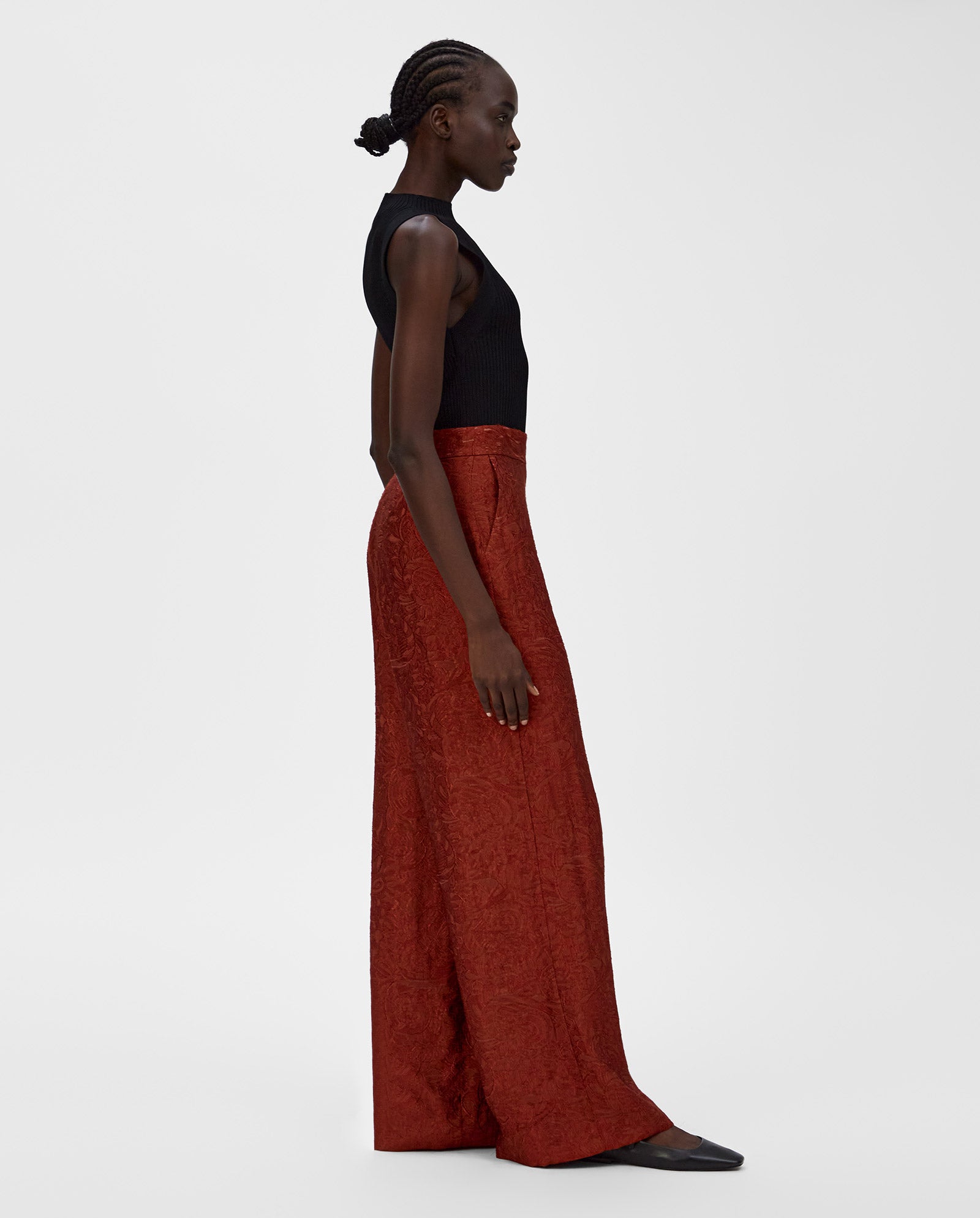 A woman in a black sleeveless top and high-waisted, wide-leg PRIYA Trousers in rust red by IVY OAK stands in profile.