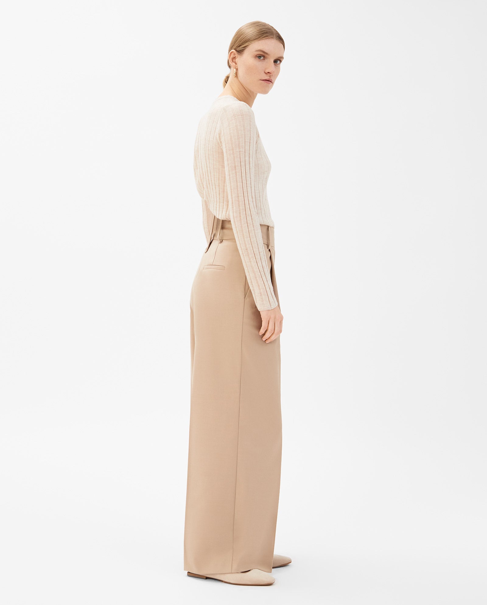 Woman in IVY OAK PRESCILLIA beige wide leg trousers, a cream ribbed sweater, and flat shoes stands sideways against a white backdrop.