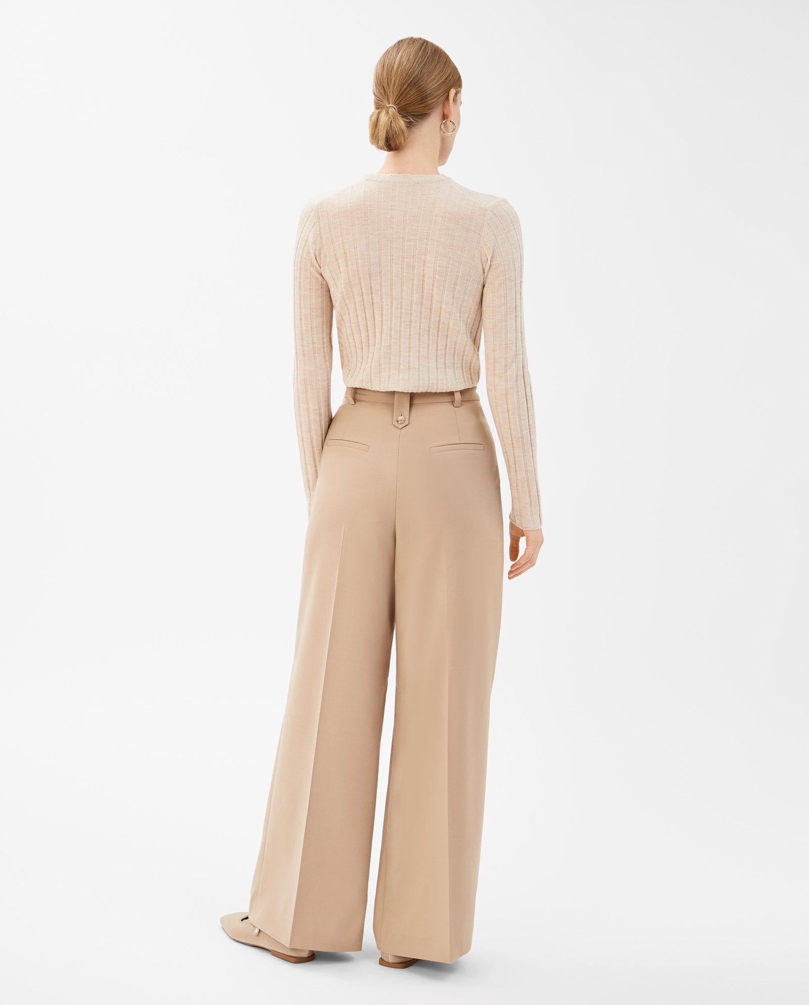 A woman stands facing away against a white background, wearing IVY OAKs PRESCILLIA Trouser in beige and a light ribbed sweater.