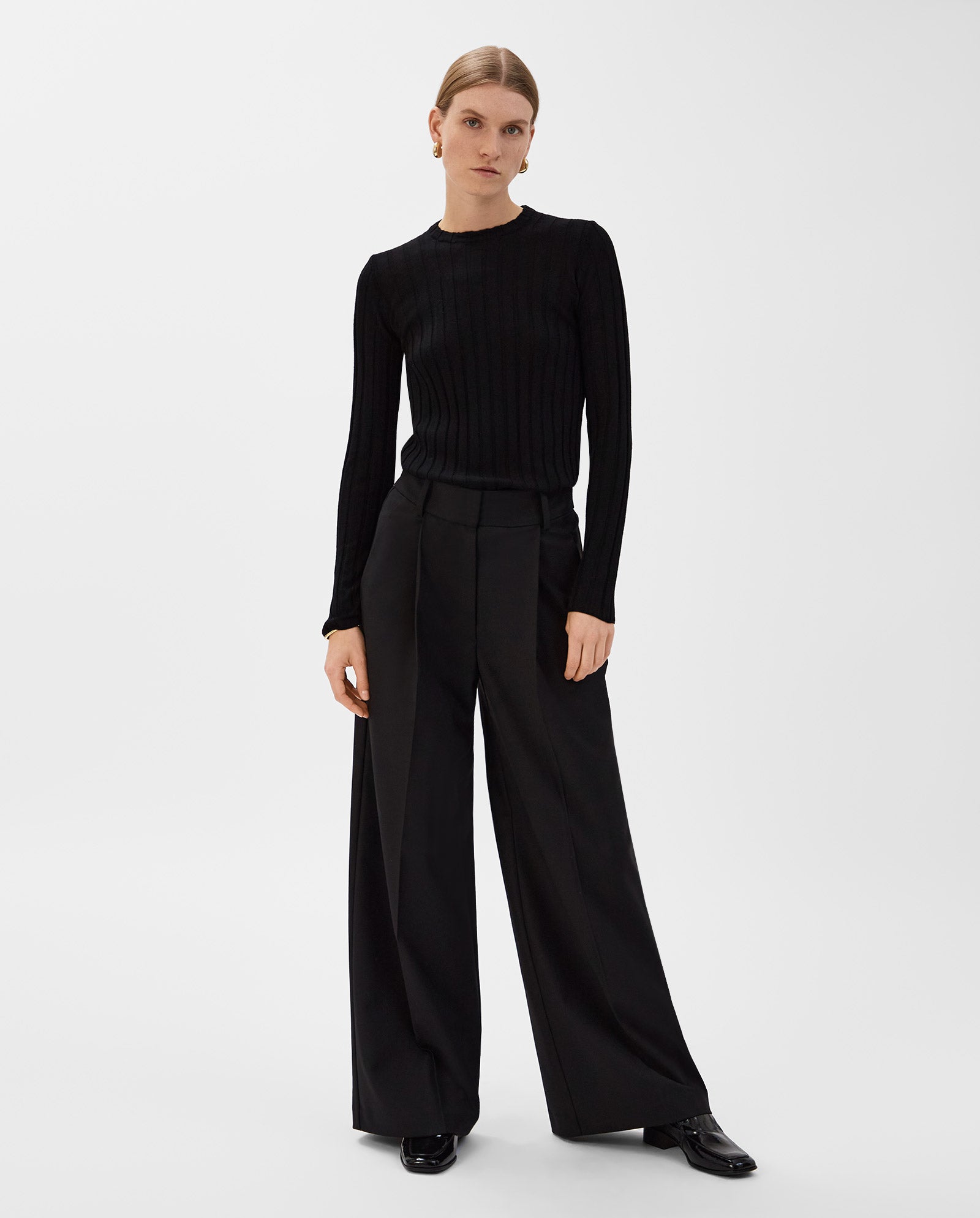 A woman stands against a white background, wearing IVY OAKs PRESCILLIA Trouser and a black ribbed sweater.