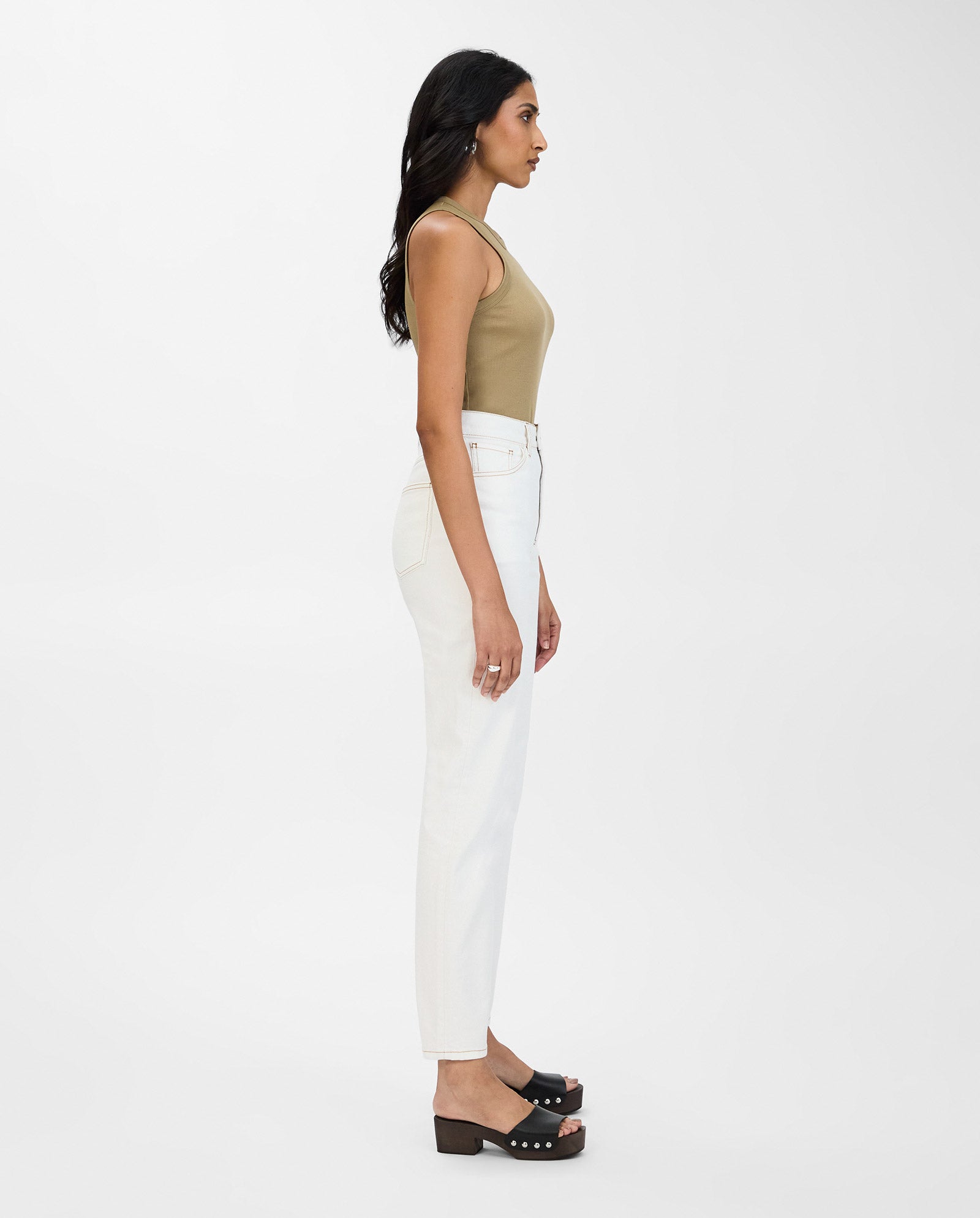 A woman in a tan sleeveless top, IVY OAKs white POEMA Trousers with a straight leg cut, and black sandals stands in profile against a white background.