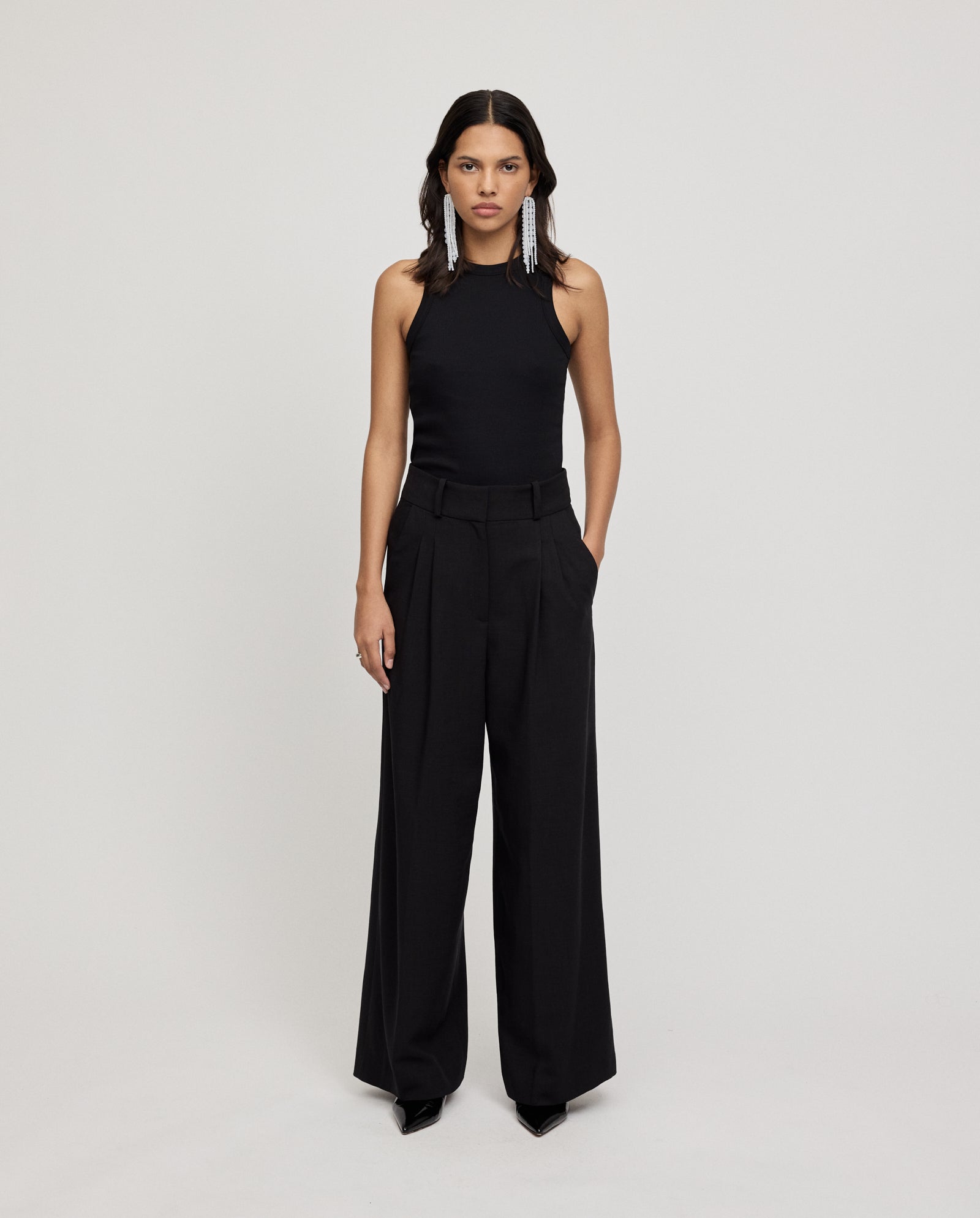 A woman wears IVY OAKs PRESCILLIA ANN Pants—high-waist classic trousers with French pockets—paired with a sleeveless black top, standing against a plain background.