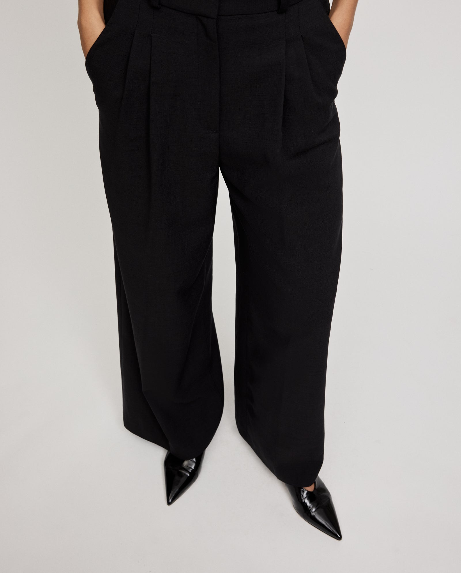 A person wears IVY OAK PRESCILLIA ANN high-waisted, wide-leg classic trousers with French pockets and black pointed-toe shoes, hands in pockets.