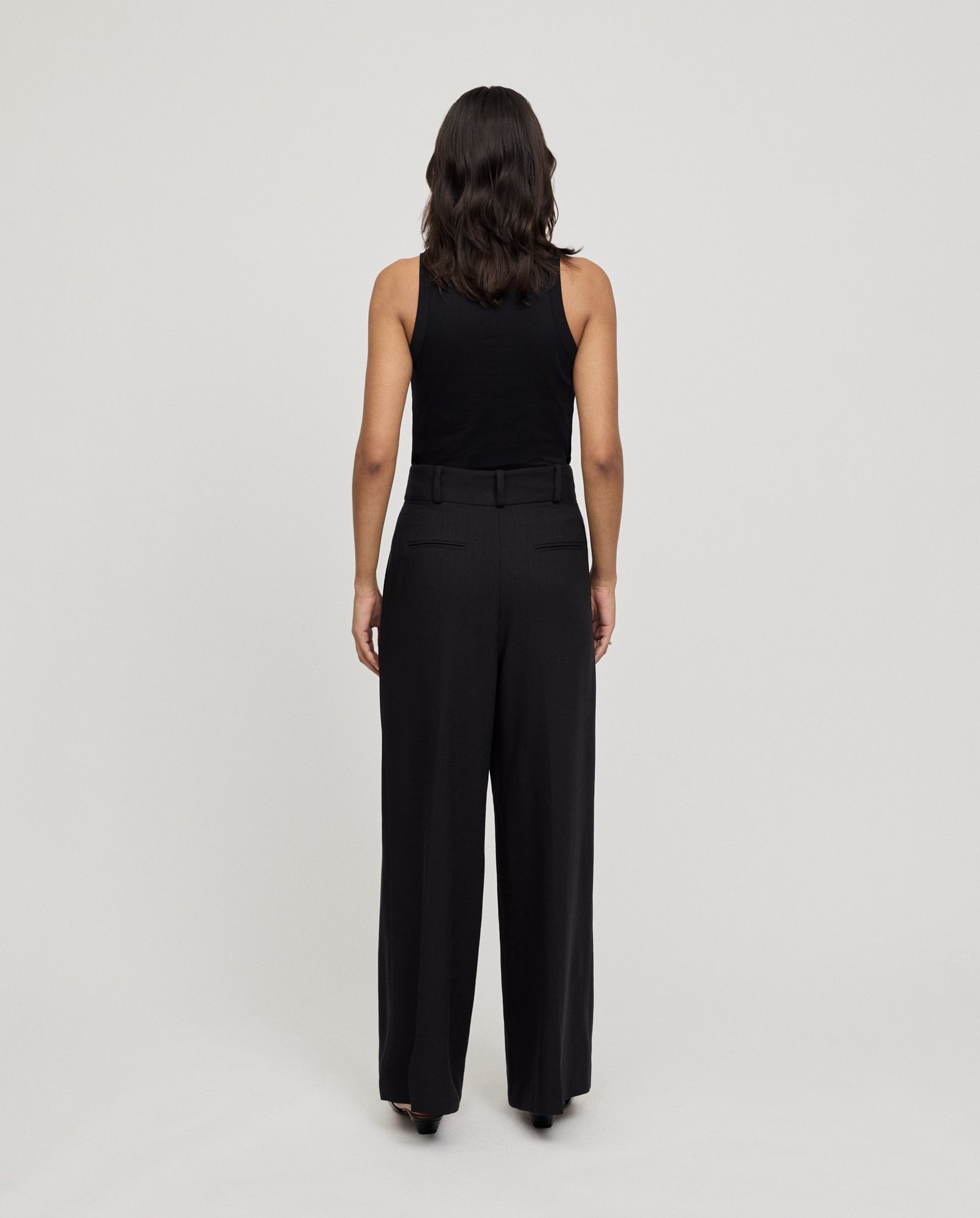 A woman faces away in a black sleeveless top and IVY OAK PRESCILLIA ANN classic high waist trousers with French pockets, set against a plain background.