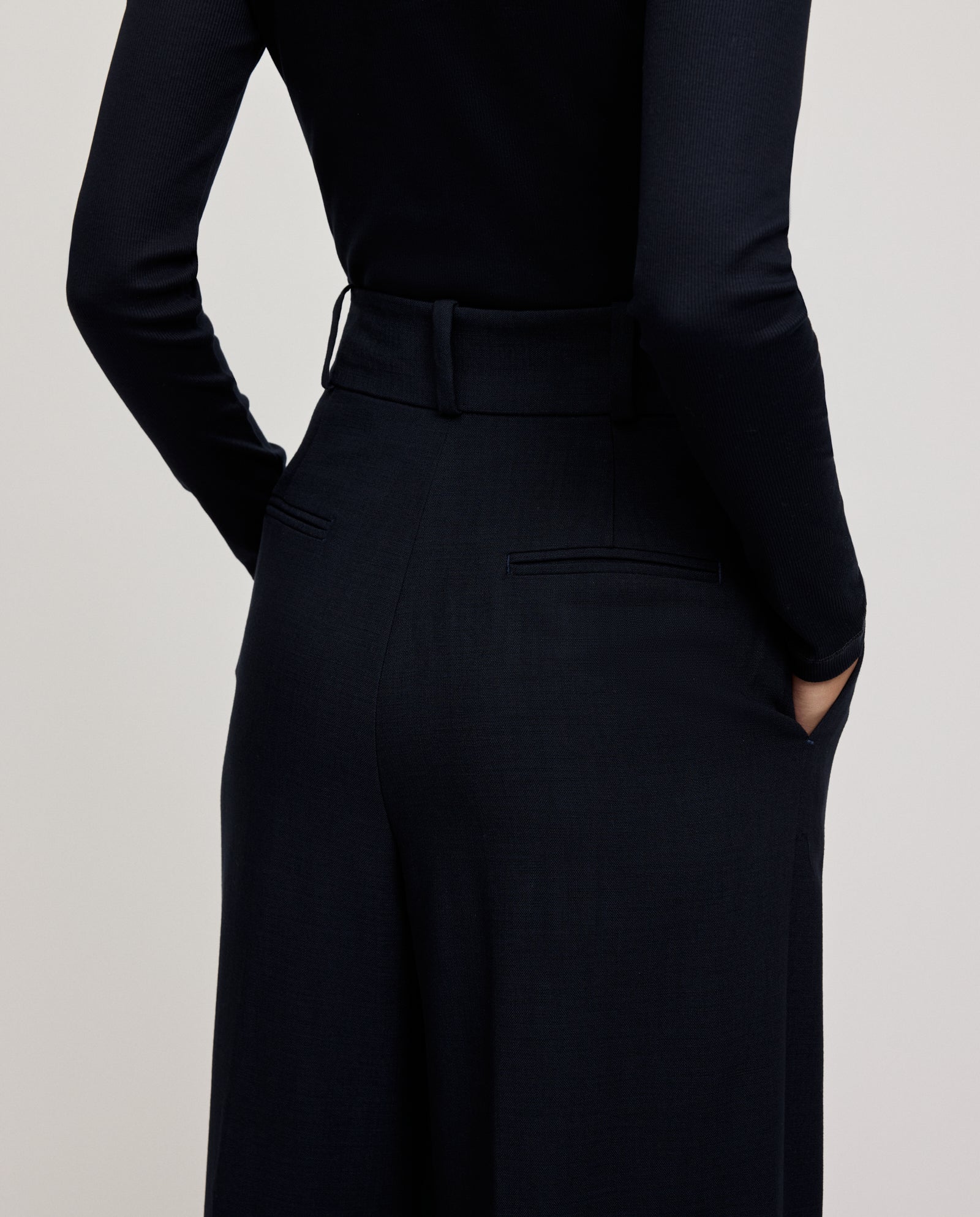 A person, viewed from behind with hands in pockets, wears IVY OAK’s PRESCILLIA ANN Pants featuring French pockets and a chic cut, styled with a black long-sleeve top.