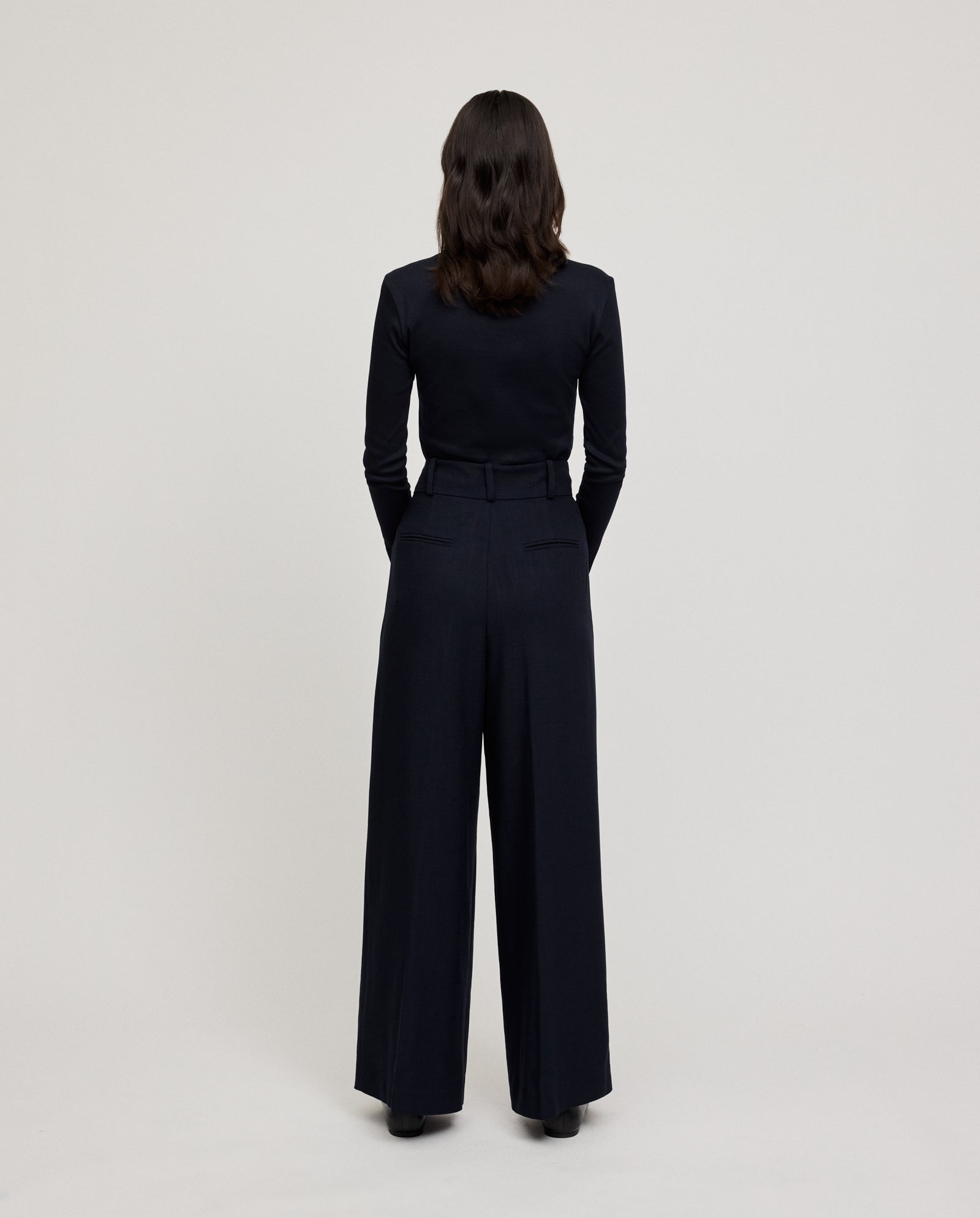 A person with shoulder-length dark hair, dressed in a black top and PRESCILLIA ANN Pants by IVY OAK, stands facing away.