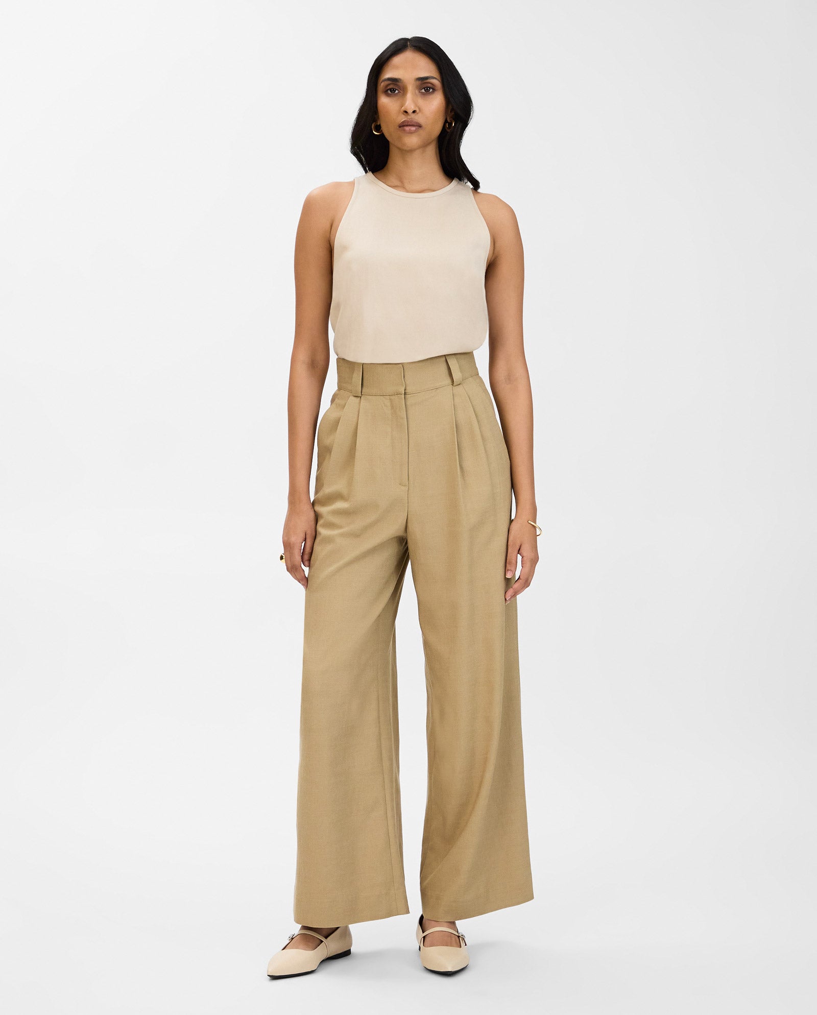 A woman wears IVY OAK PRESCILLIA ANN high-waisted, wide-leg pleated trousers with a beige sleeveless top and matching beige flats.