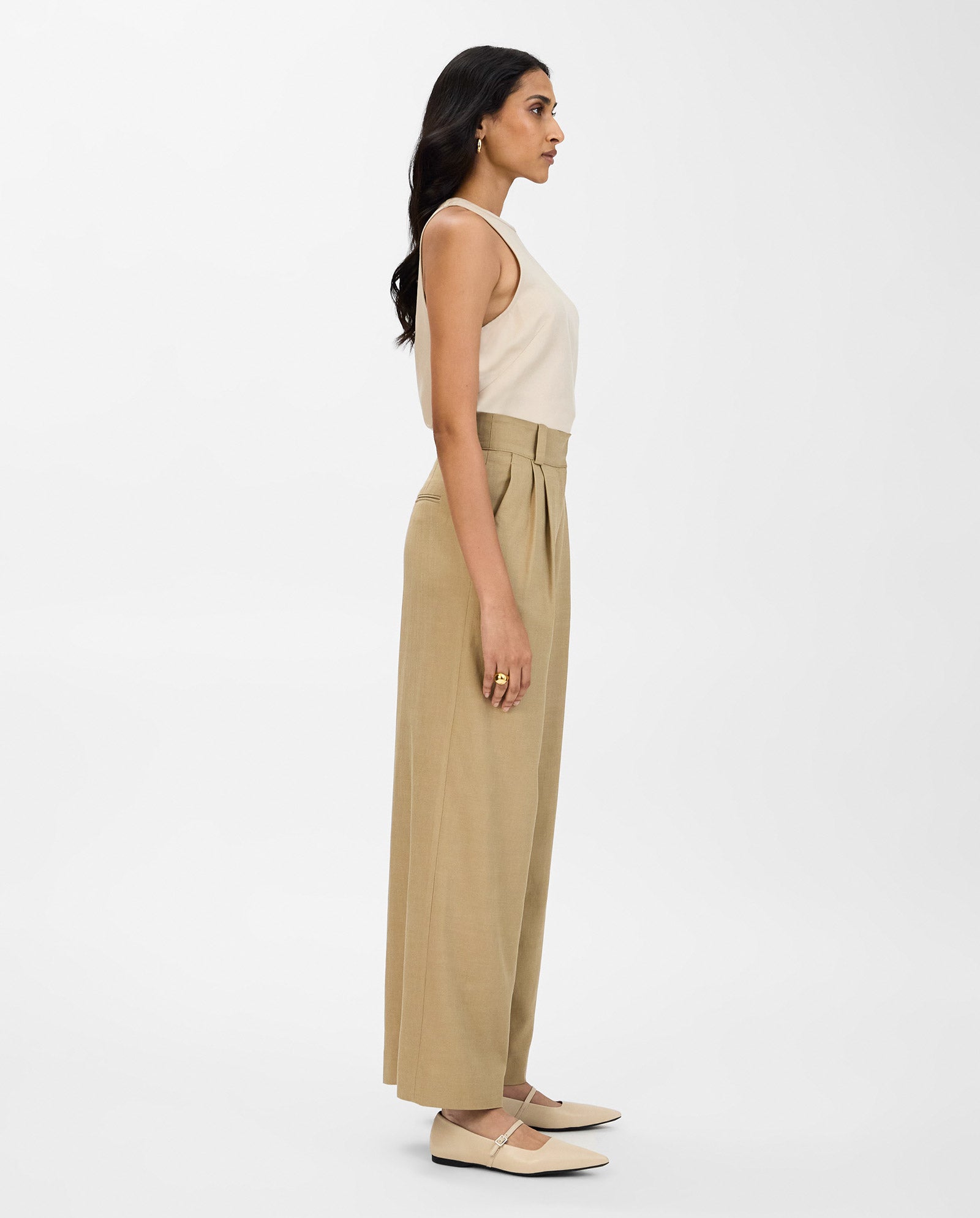 Woman in beige PRESCILLIA ANN wide leg trousers with soft pleats by IVY OAK and a sleeveless top, standing in profile against a plain white background.