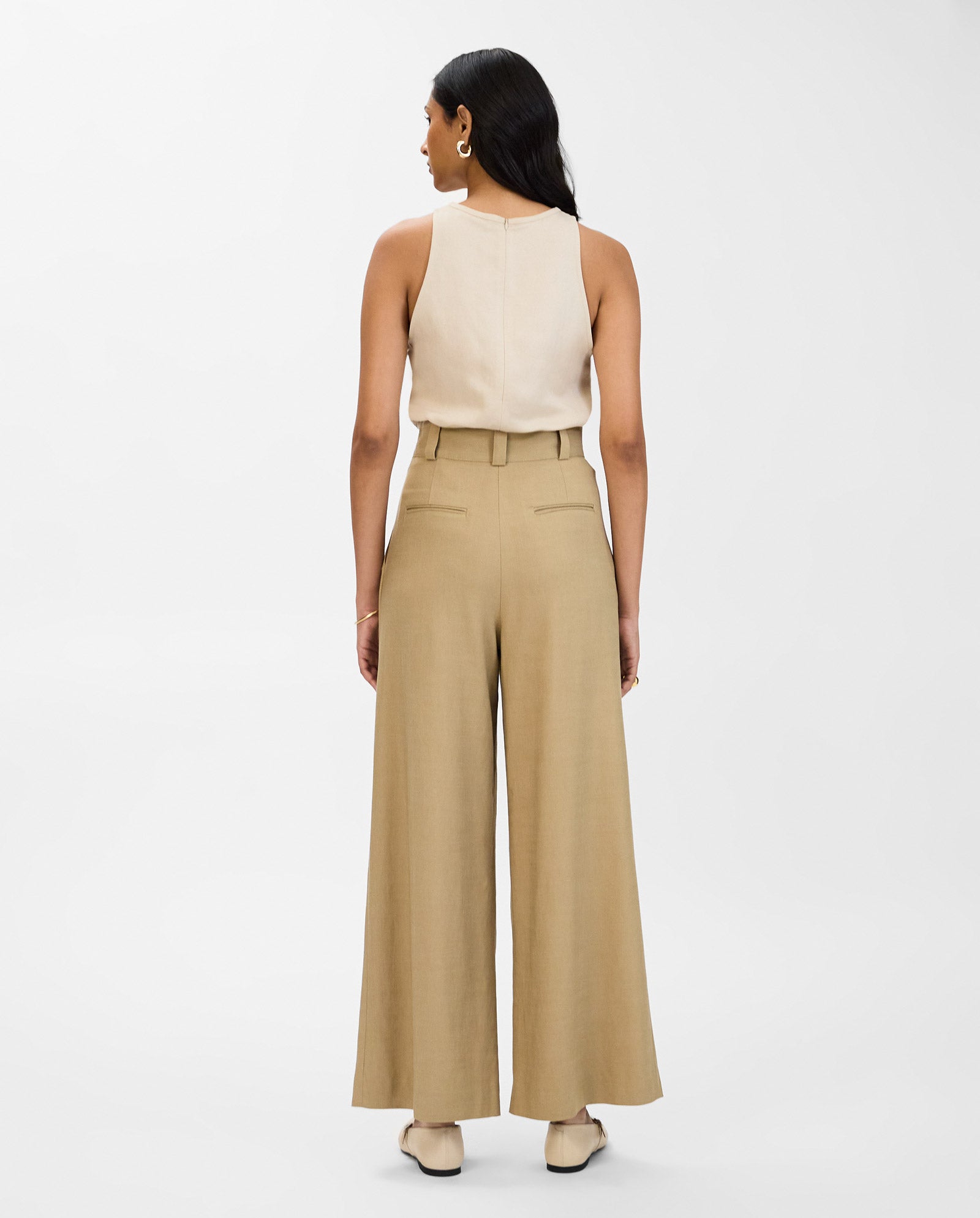 A woman faces away, wearing IVY OAK PRESCILLIA ANN wide-leg beige trousers with subtle pleats, a sleeveless top, and beige shoes.