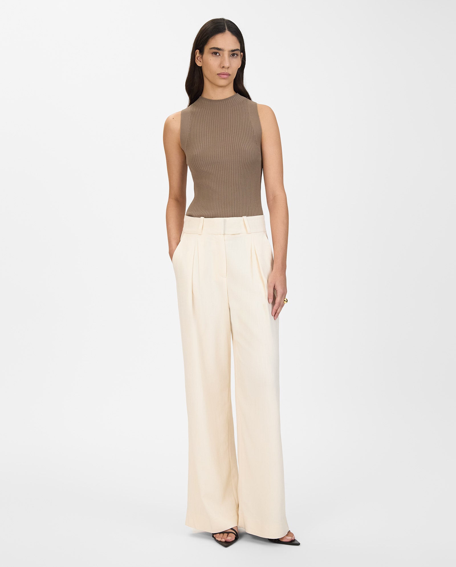 A woman stands with hands in pockets, wearing IVY OAK’s PRESCILLIA ANN Pants—high-waisted, wide-leg trousers—paired with a sleeveless brown top.