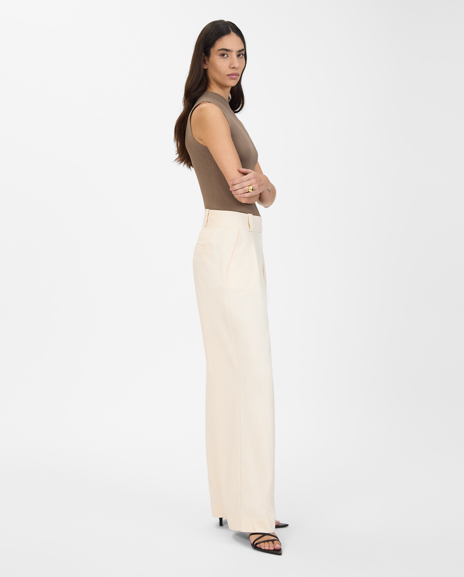 A woman stands sideways with arms crossed, wearing IVY OAKs PRESCILLIA ANN Pants—cream wide leg trousers—paired with a sleeveless brown top.
