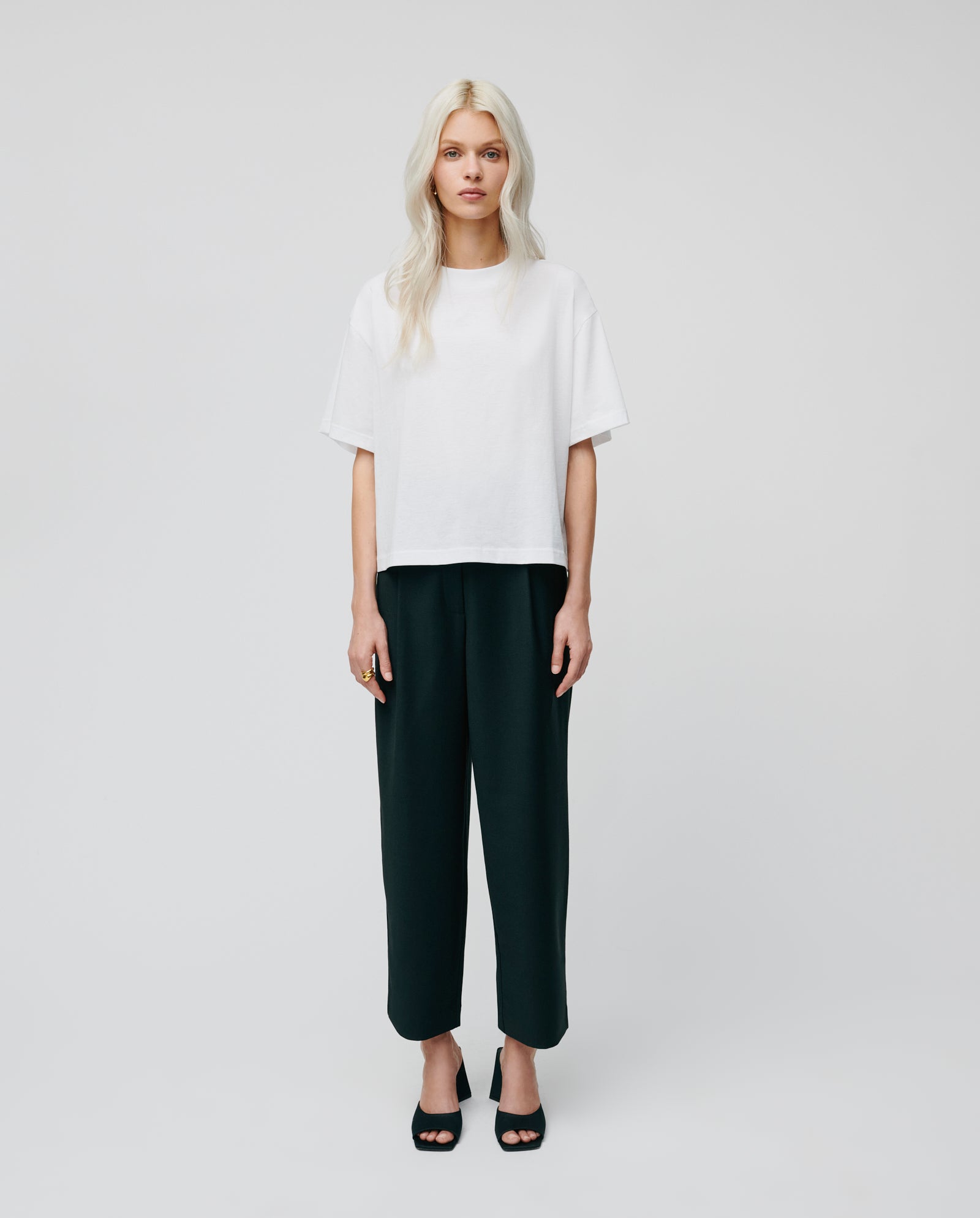 A woman with long blonde hair wears a white t-shirt, black IVY OAK PERSEPHONE ANN Trousers with a relaxed tapered cut, and black heels, standing against a plain background.