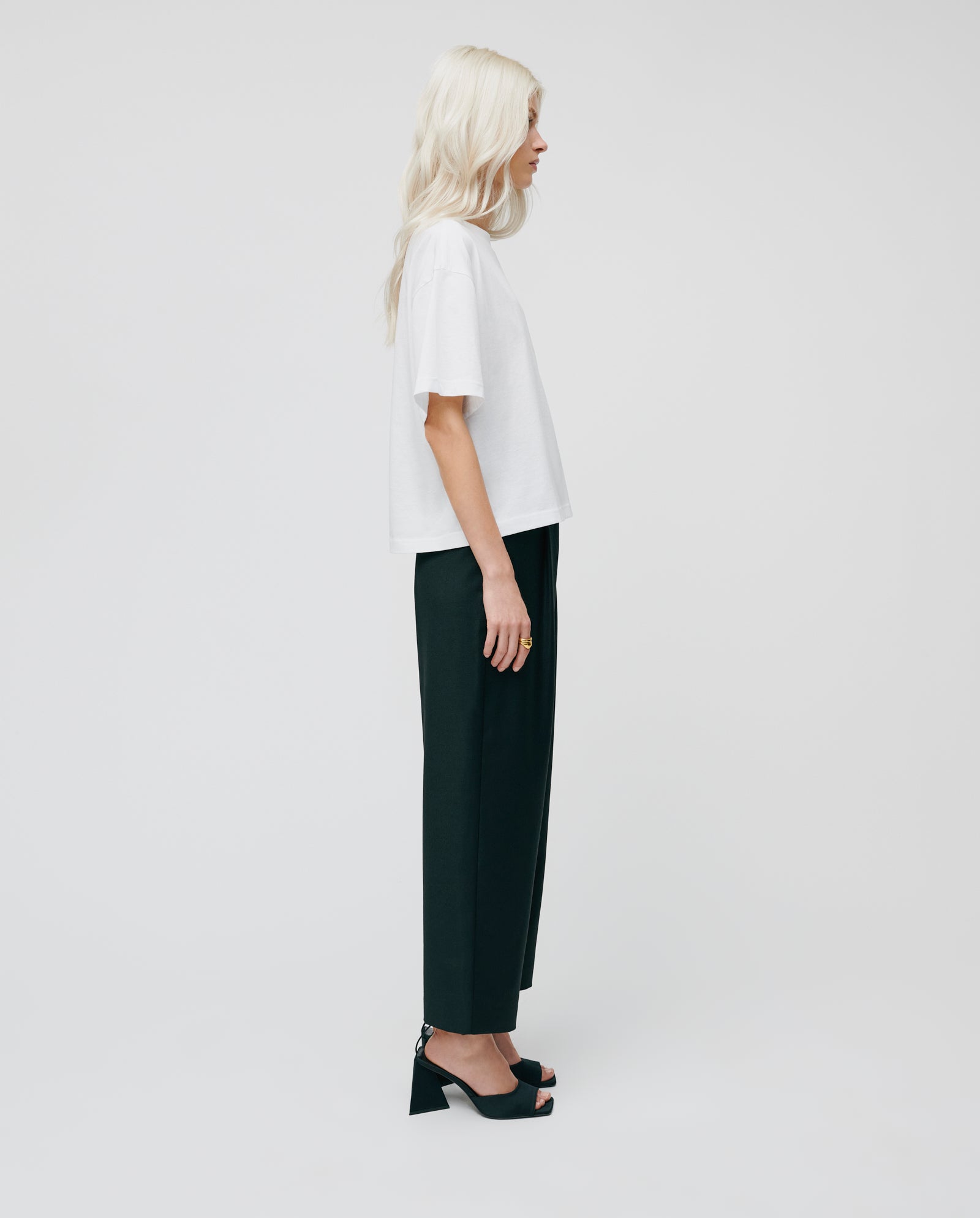 A woman with long blonde hair wears a white t-shirt, IVY OAK PERSEPHONE ANN Trousers with a tapered leg, and heels, standing in profile against a white background.