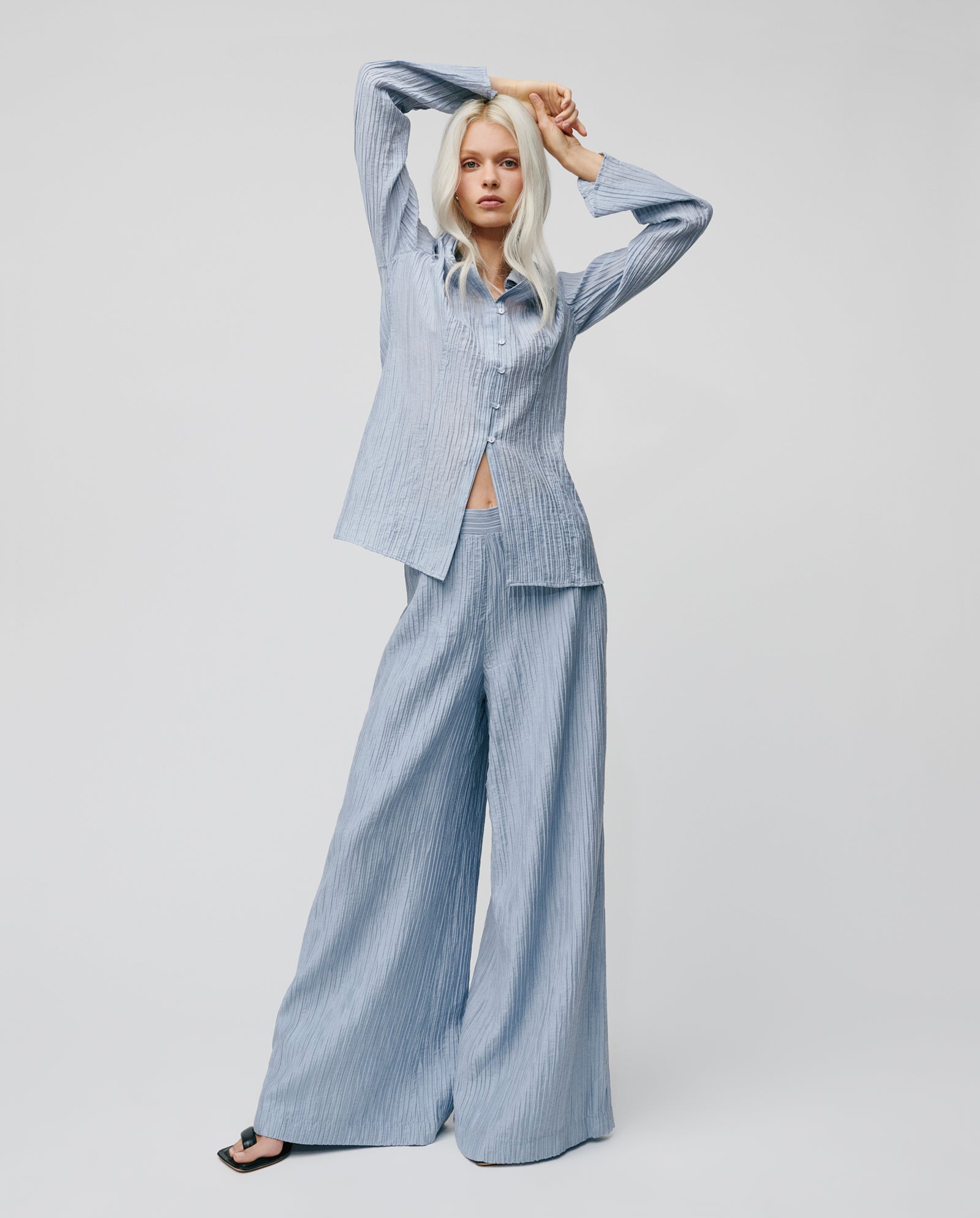 Woman wearing wide-leg pleated PORTIA Trousers by IVY OAK and a loose, light blue pinstriped shirt poses with arms raised above her head.