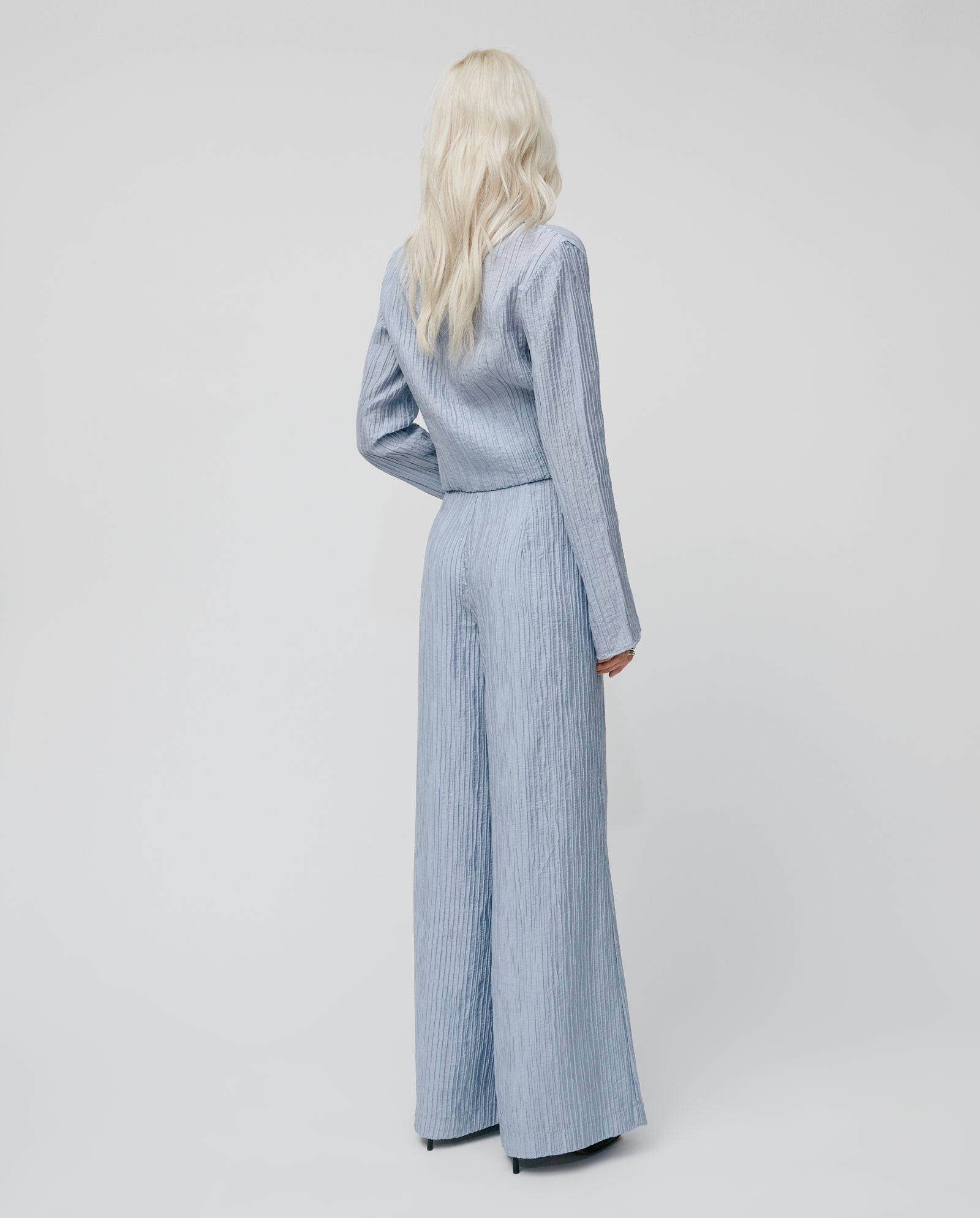 A person with long blonde hair stands facing away from the camera, wearing IVY OAKs PORTIA Trouser in light blue with pleated detailing.