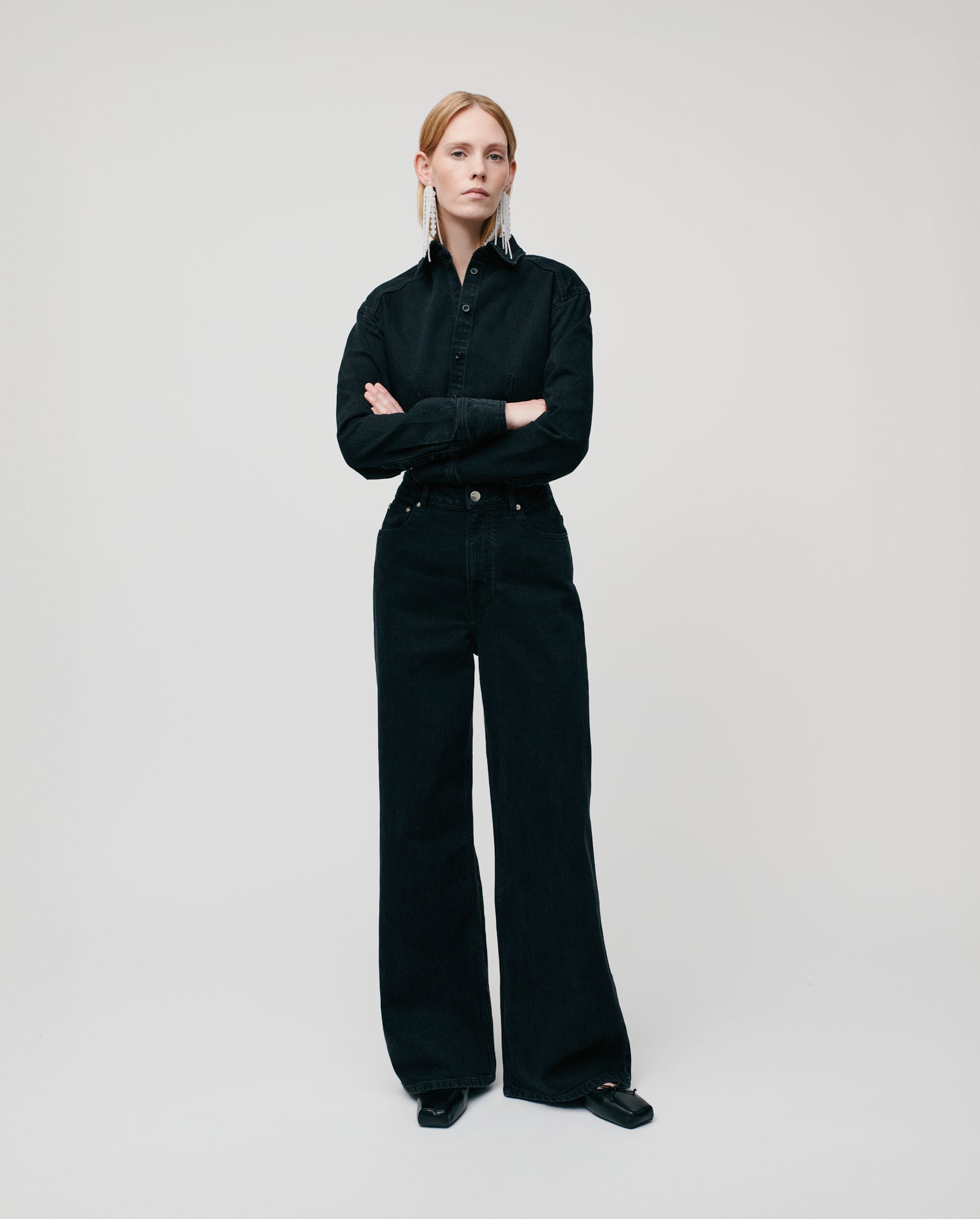 Person standing with arms crossed, wearing a black shirt and IVY OAK PRIJANKA Trousers in an indigo wide-leg silhouette against a plain background.